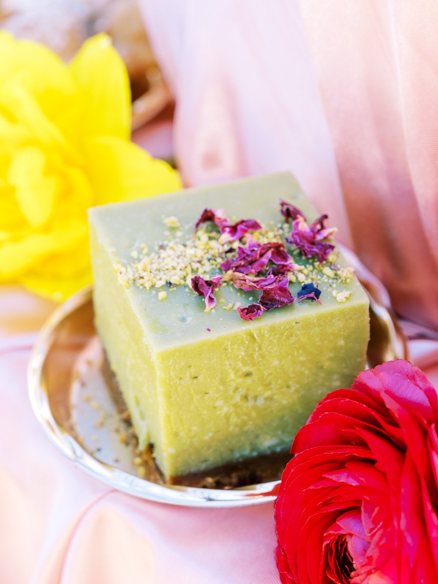 Green pistachio dessert topped with edible flowers beside vibrant ranunculus blooms on blush satin wedding display.