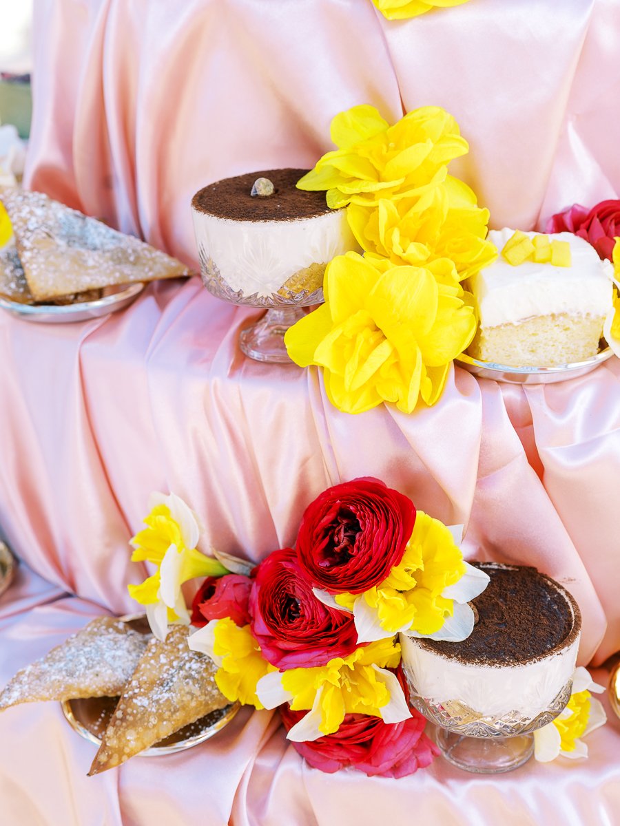 Dessert display with tiramisu, pastries, and bright floral arrangements styled on blush satin fabric for wedding reception.