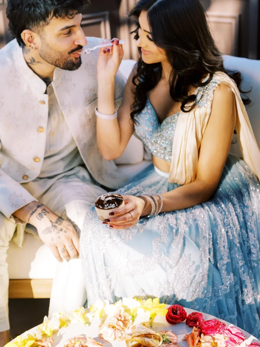 South Asian couple sharing dessert while seated together in pastel reception attire during wedding celebration.