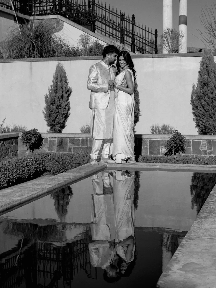 Black-and-white portrait of South Asian couple reflected in courtyard pool beside stone architecture.