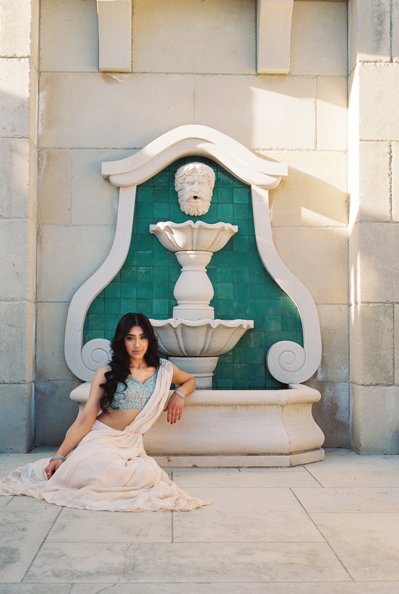 Bridal portrait in pastel blue blouse and draped sari seated beside ornate stone fountain outdoors.