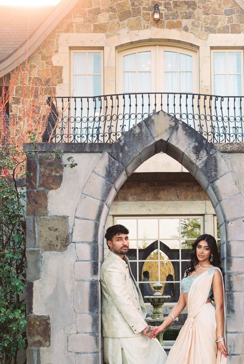 South Asian couple standing beneath stone archway in coordinated pastel wedding reception attire.
