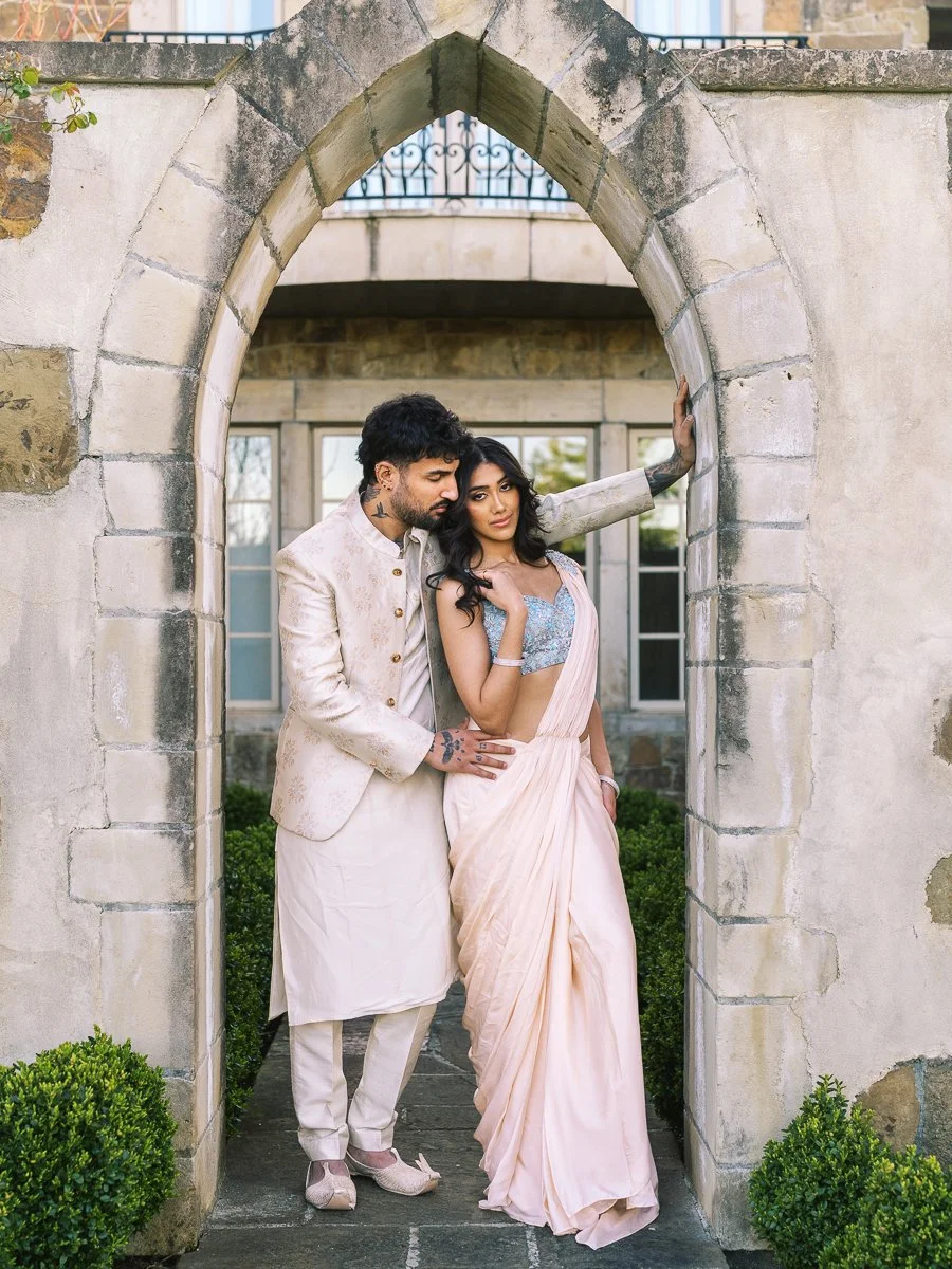 Romantic portrait of South Asian couple framed beneath stone archway in elegant outdoor courtyard setting.