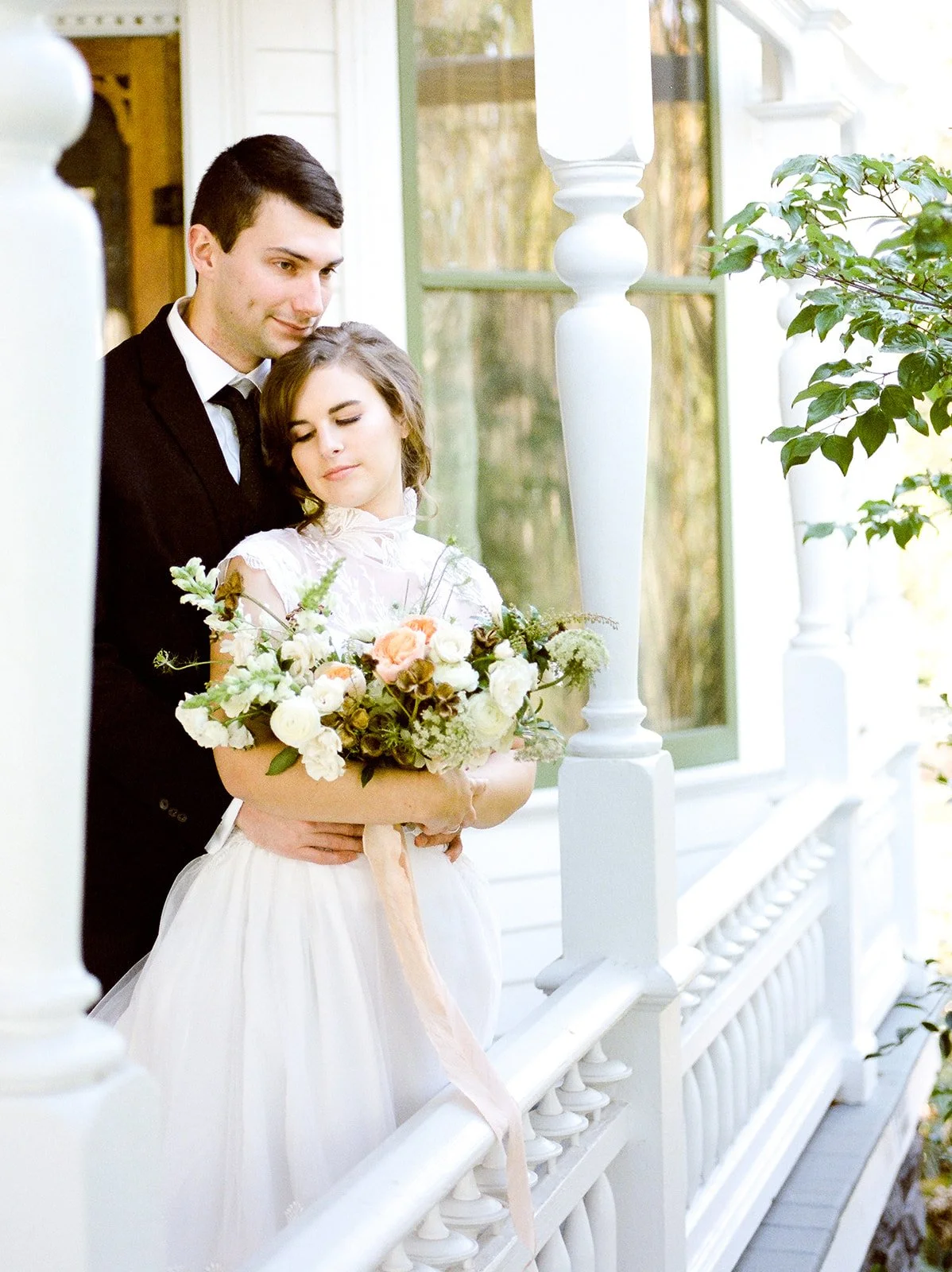 Bride holding a soft floral bouquet as the groom embraces her on the porch at Deepwood Museum &amp; Gardens.