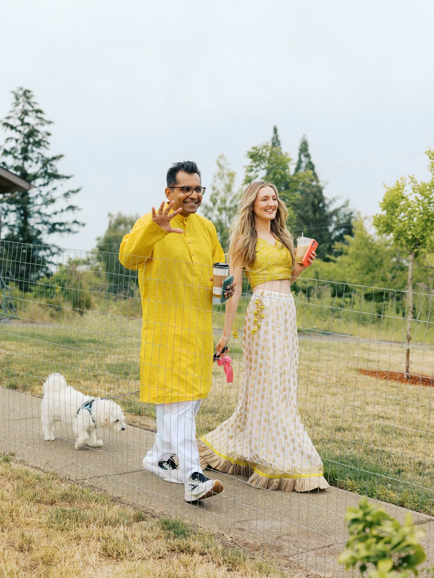 Couple in yellow Haldi attire walking outdoors with their small white dog.