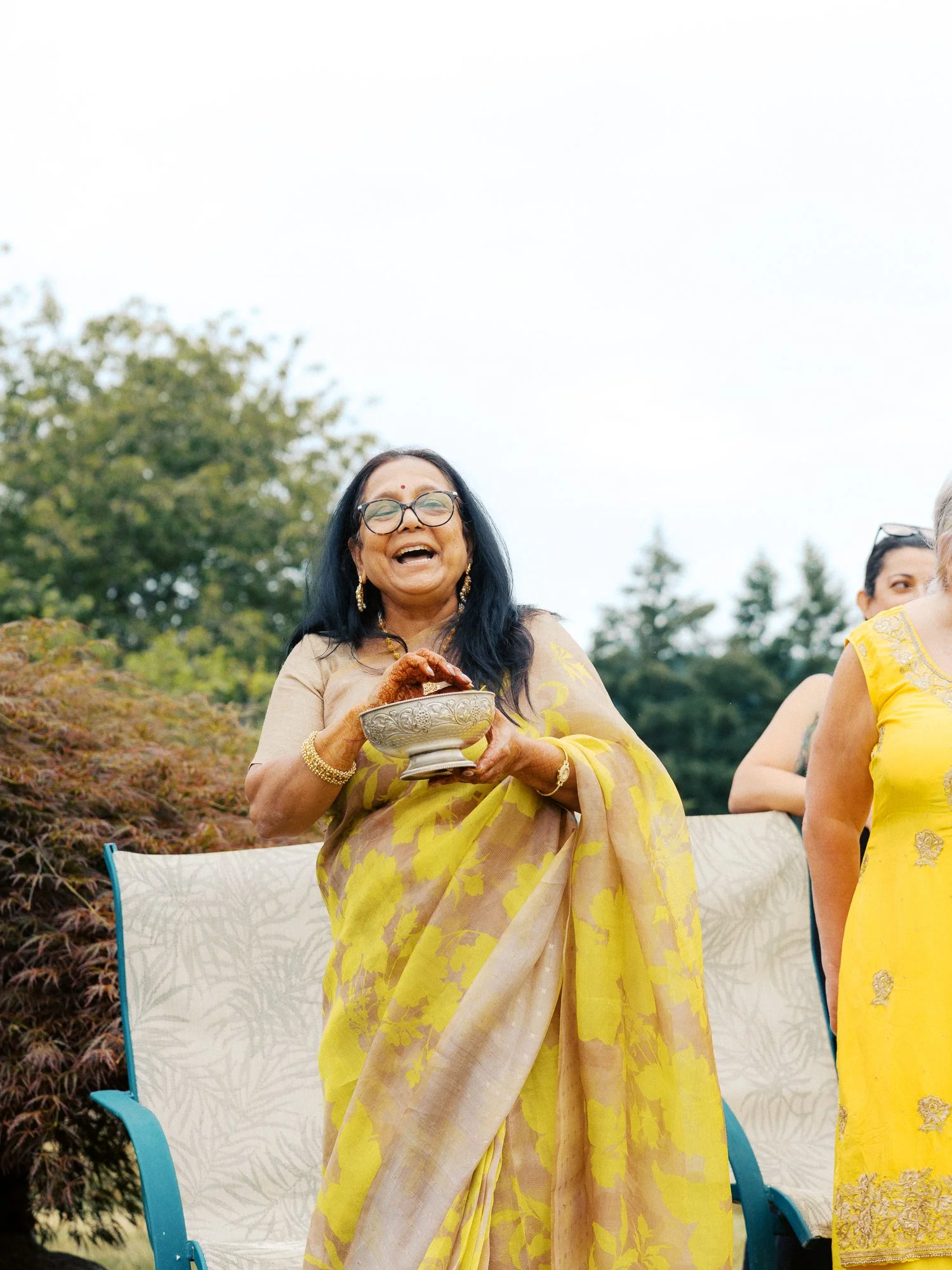Guest in a yellow floral sari laughing while holding a decorative bowl at the Haldi ceremony.