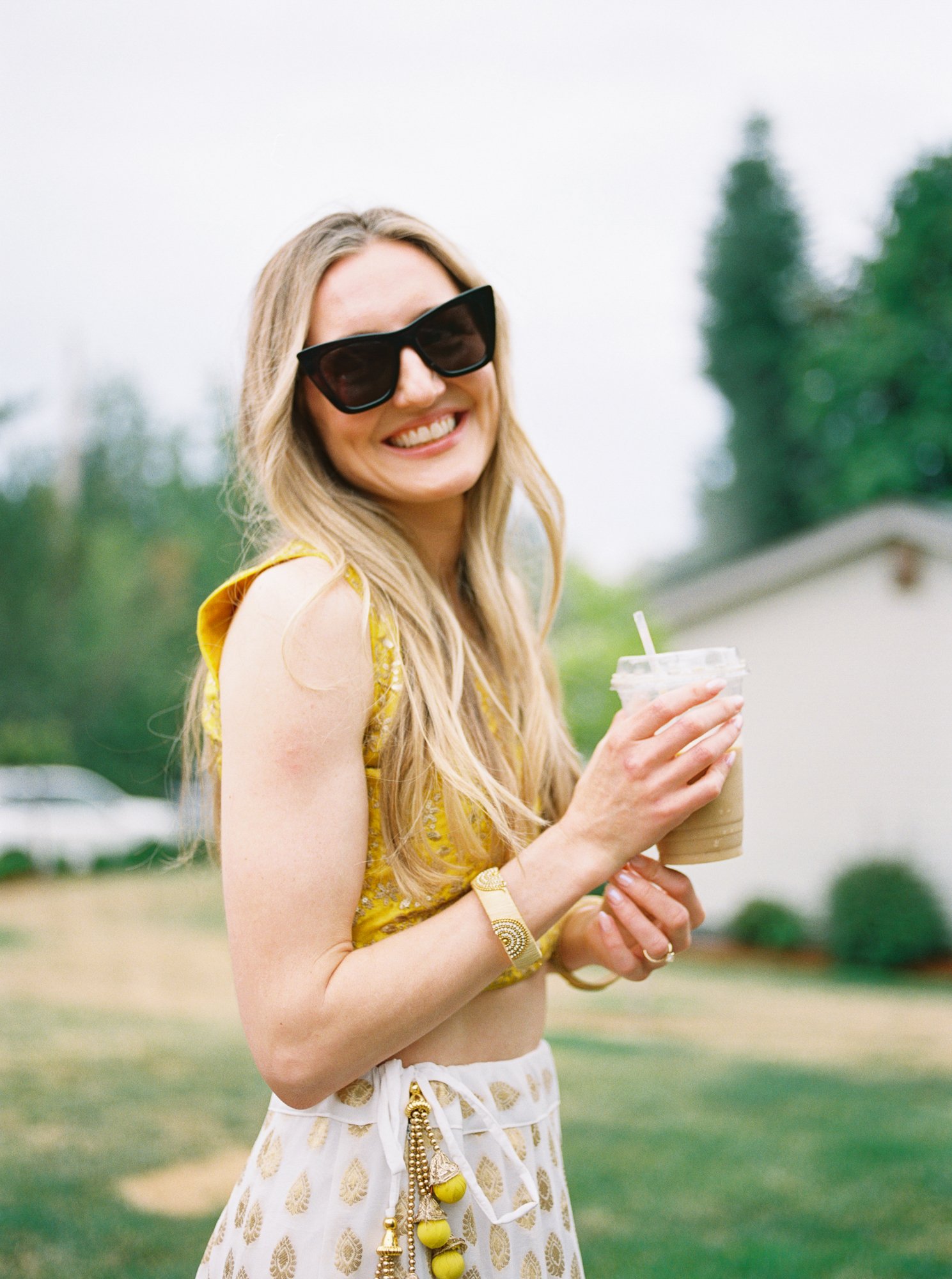 Bride in yellow and white Haldi attire smiling outdoors while holding a drink.