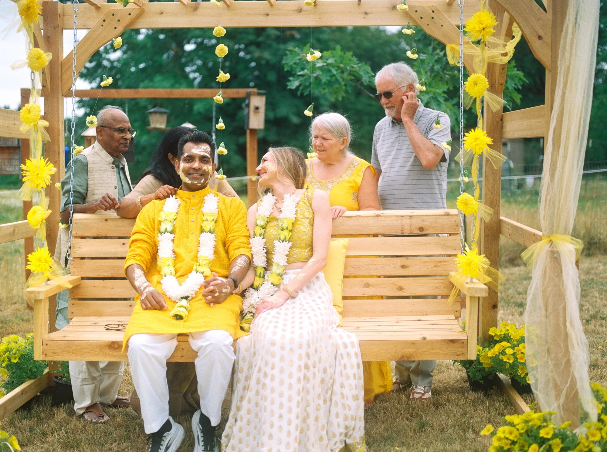 Couple seated on a flower-decorated wooden swing with family gathered behind them during Haldi.