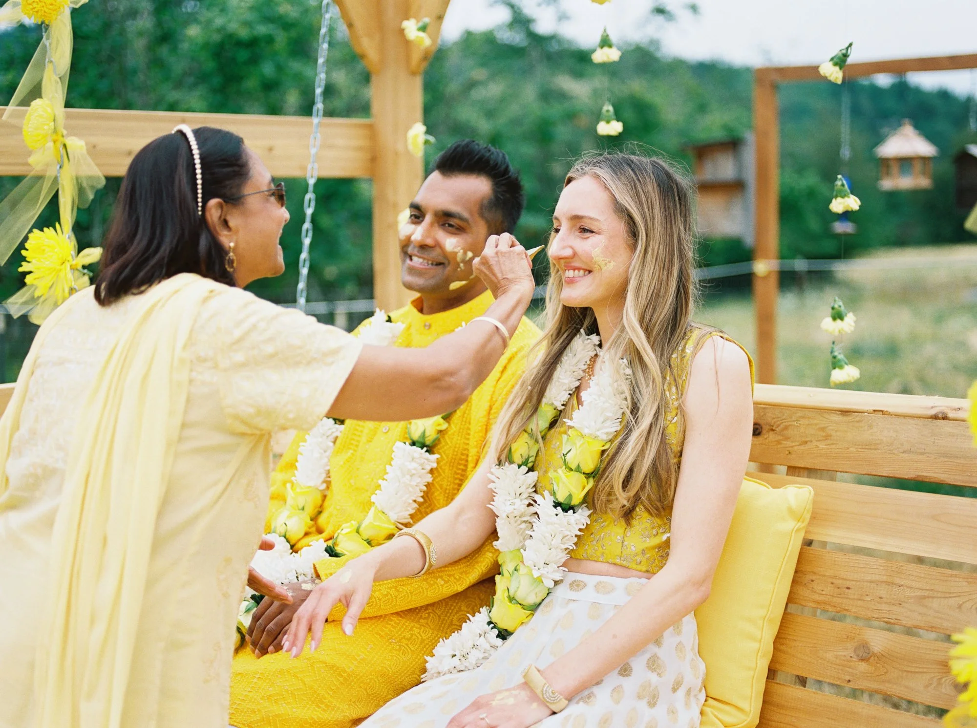 Guest applies Haldi paste to the bride’s cheek as the groom watches beside her.
