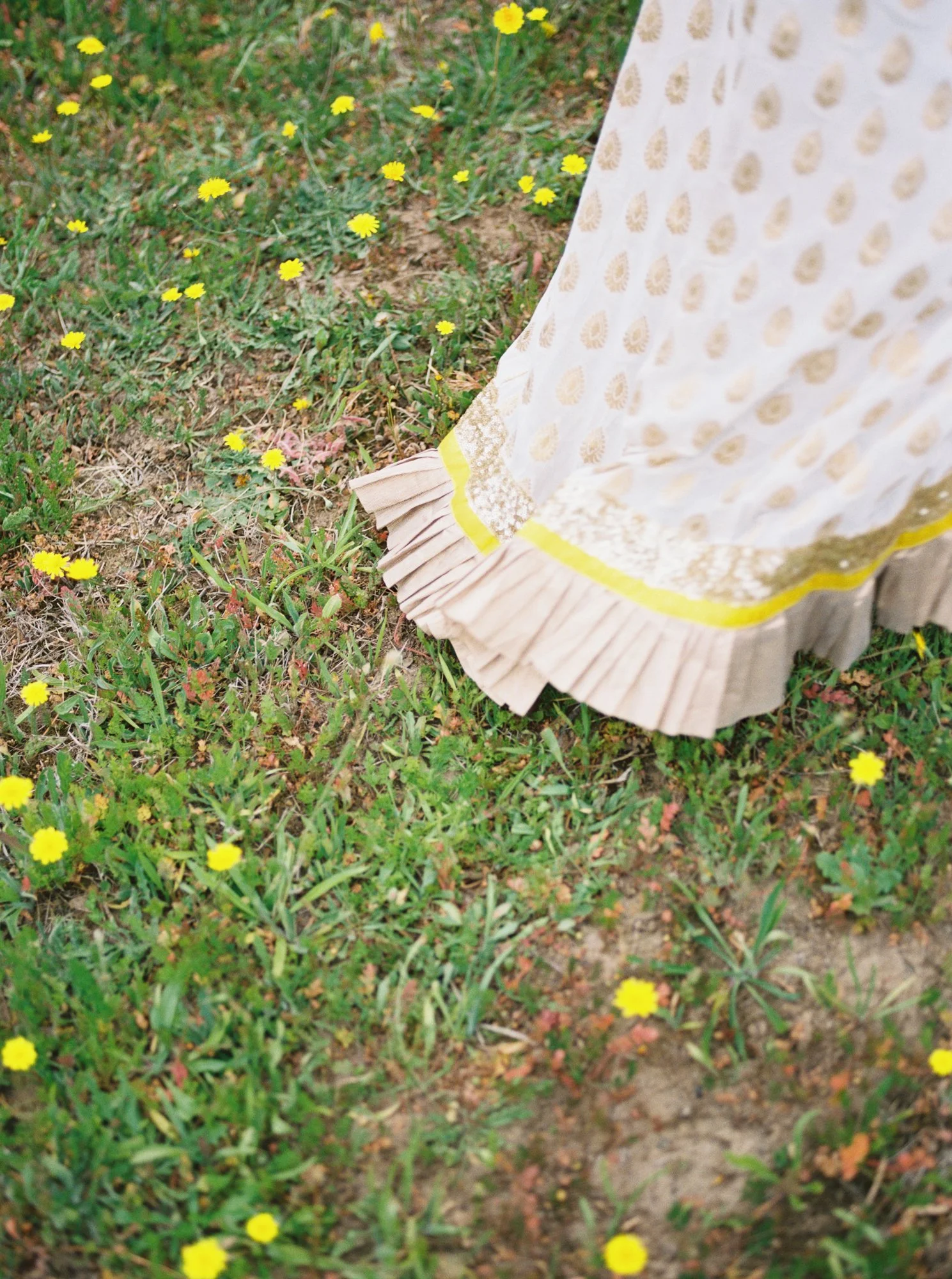 Close-up of Alacia’s white and gold Haldi skirt beside small yellow wildflowers in the grass.