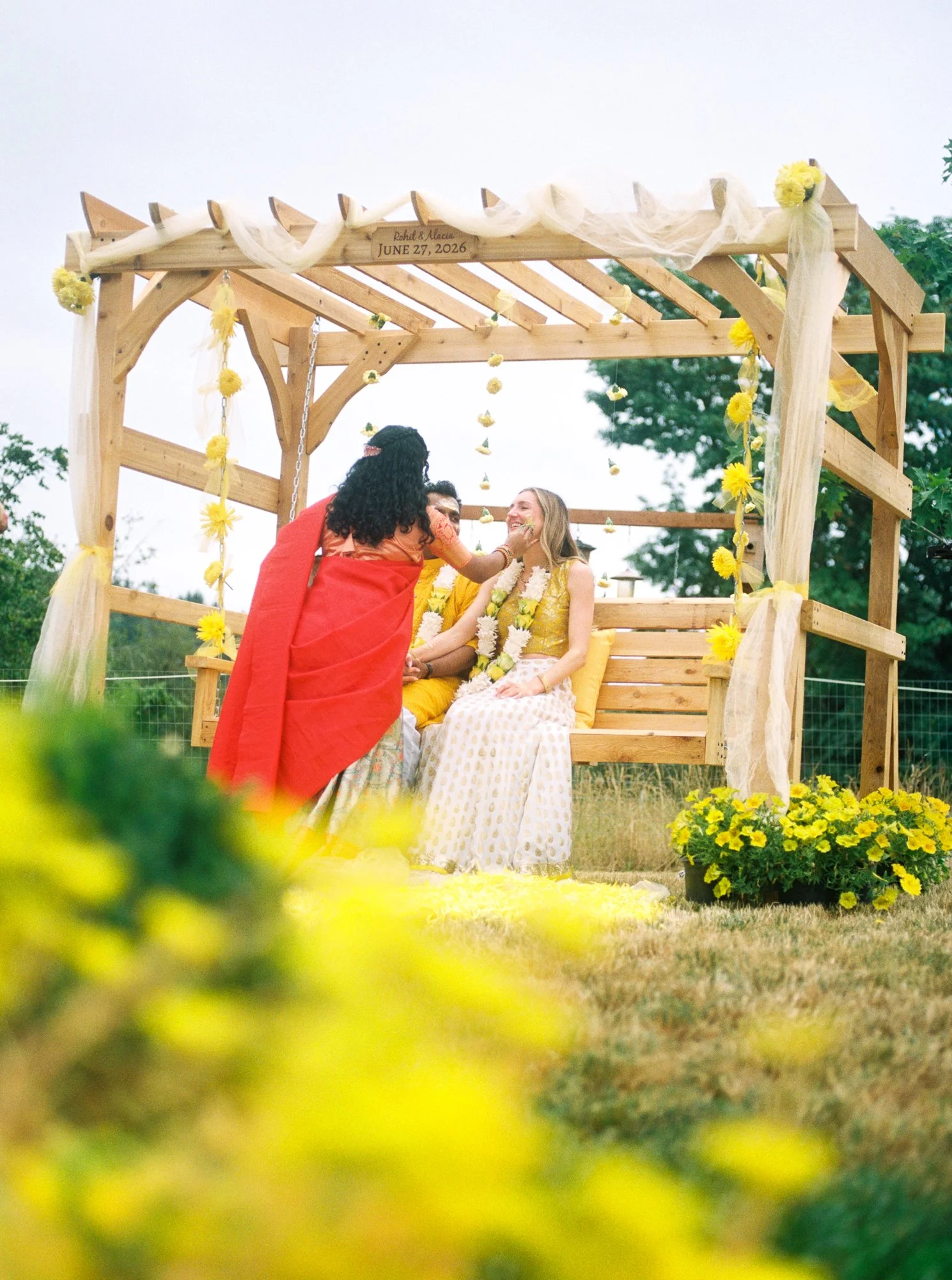 Guest applies Haldi paste to the bride while the couple sits beneath a yellow floral pergola.