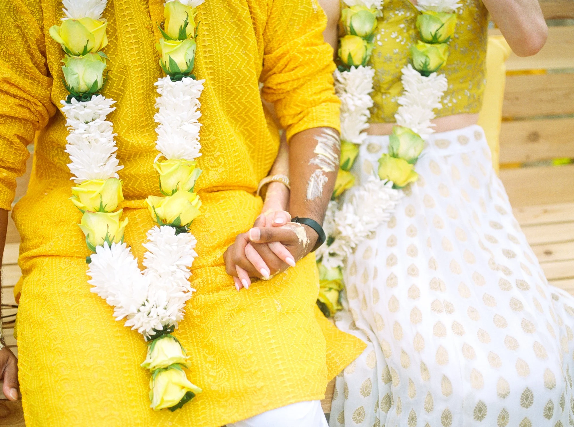Close-up of the couple holding hands while wearing yellow and white flower garlands during Haldi.