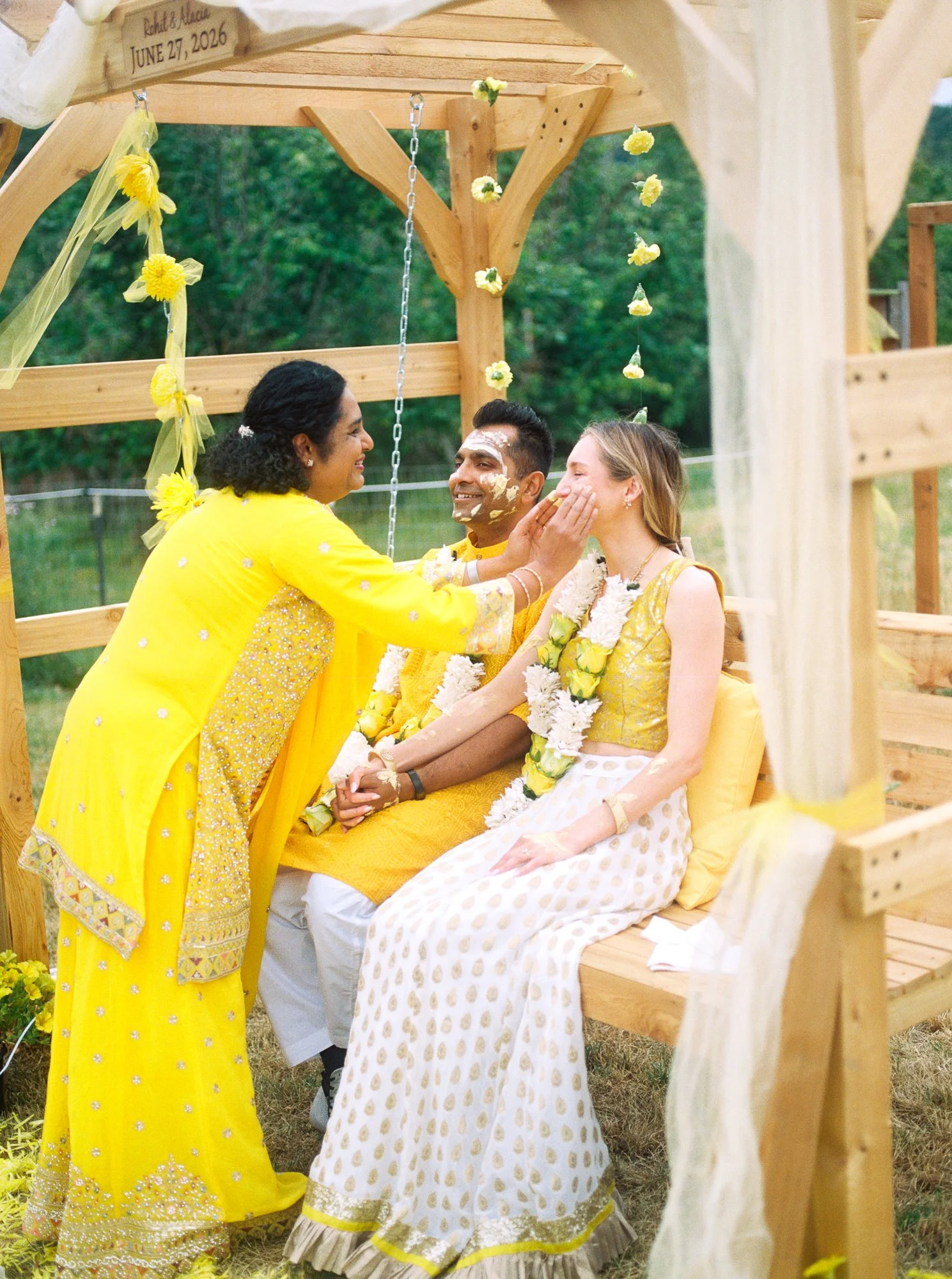 Guest applies Haldi paste to the bride’s cheek as the groom watches beside her.