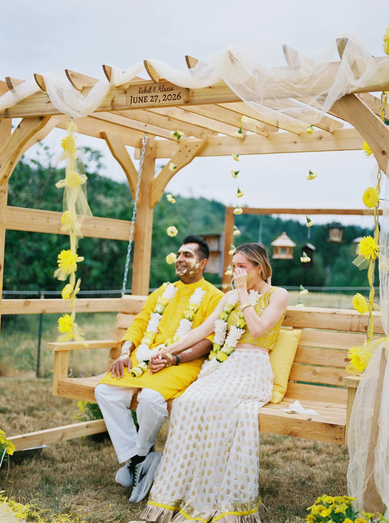 Alacia and Rohit seated together beneath a flower-decorated wooden pergola during their outdoor Haldi ceremony.