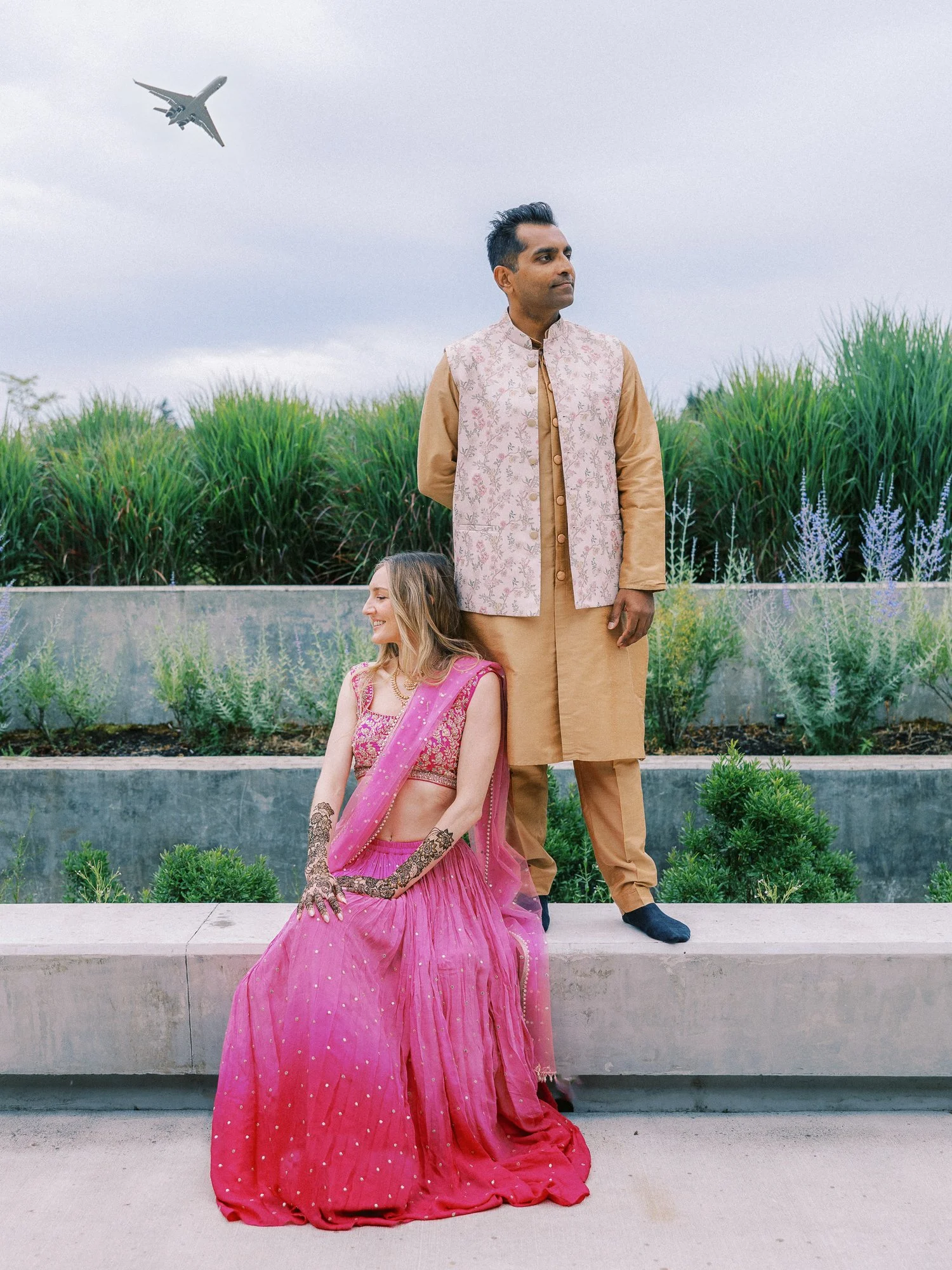 Alacia and Rohit posing outdoors in pink and beige traditional attire as an airplane passes overhead.