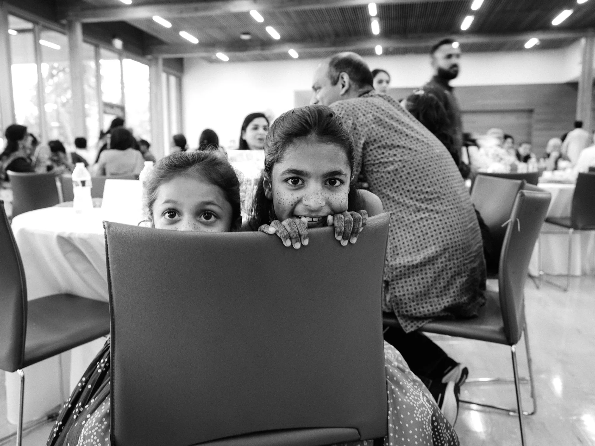 Black-and-white candid of two young girls peeking over a chair during the Mehndi reception.