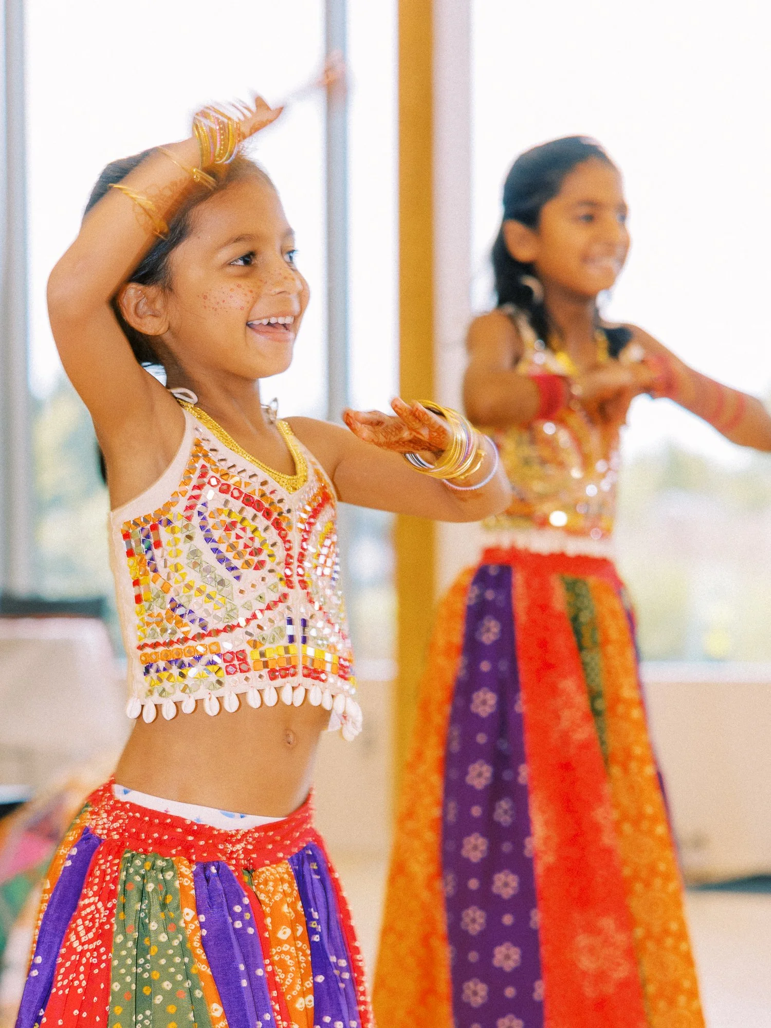 Two young girls dance in matching colorful outfits in front of the floral Mehndi stage.
