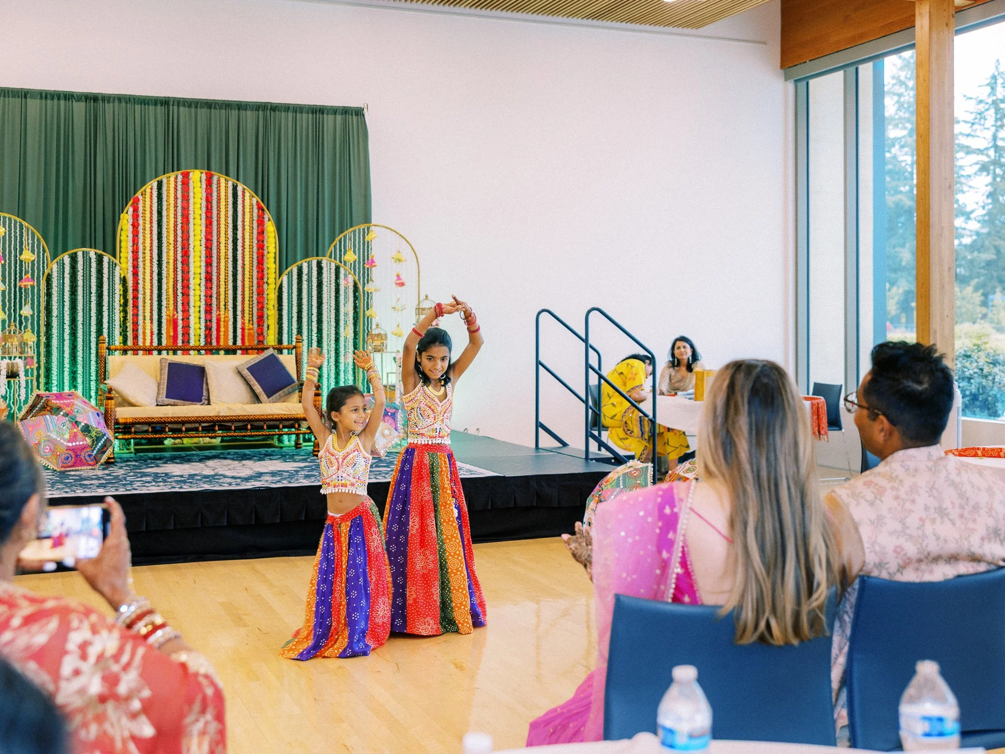 Two young girls dance in matching colorful outfits in front of the floral Mehndi stage.