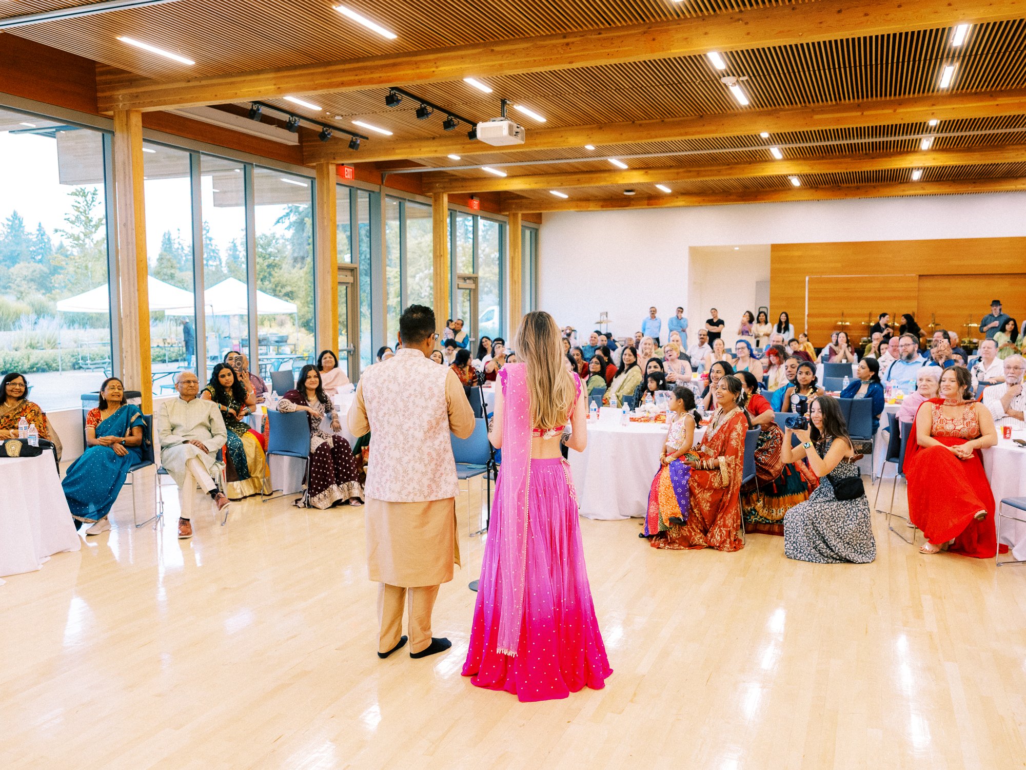 Alacia and Rohit stand together facing guests during their indoor Mehndi celebration.