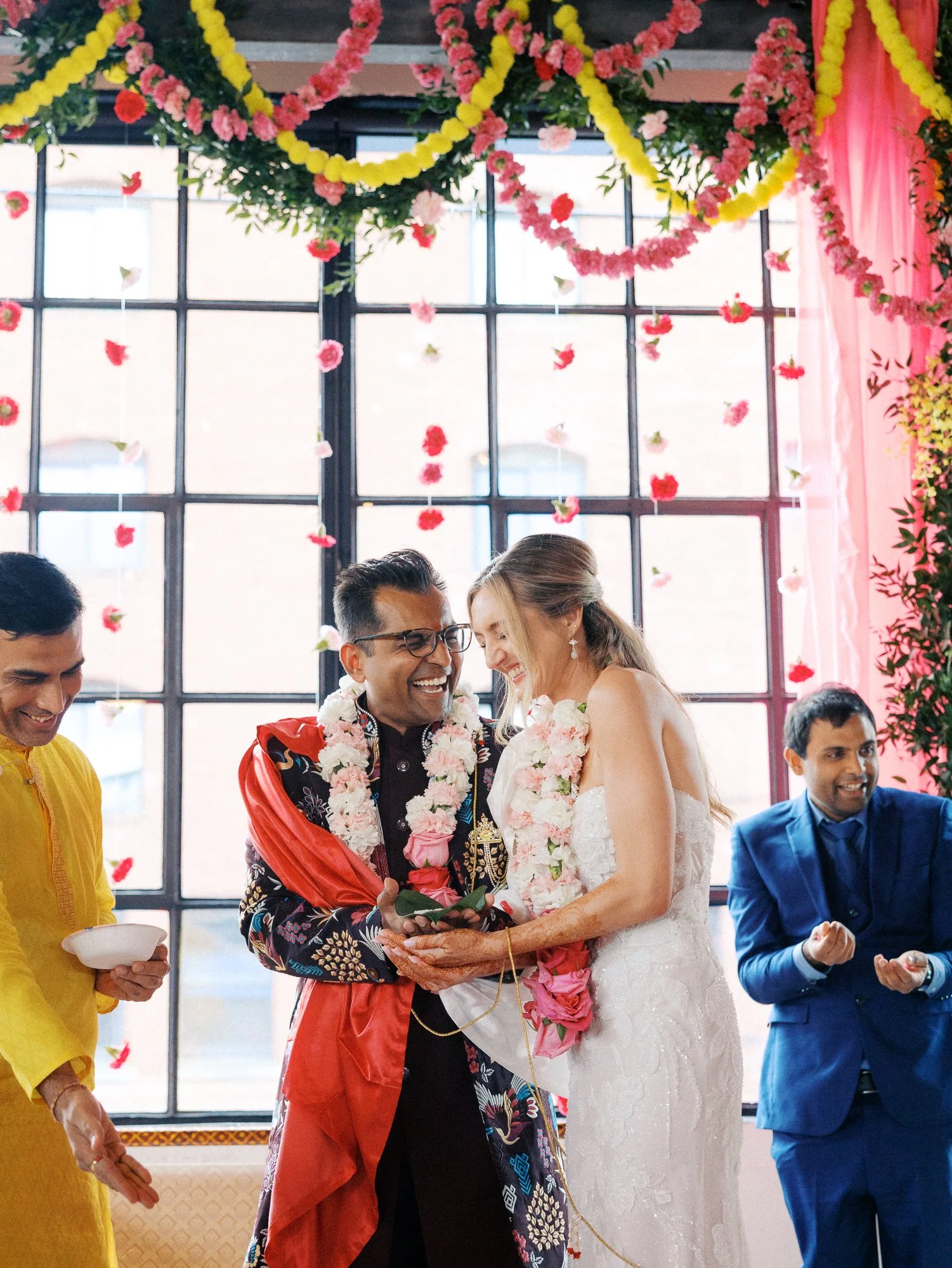 Bride and groom laugh together beneath pink and yellow floral garlands during their ceremony.