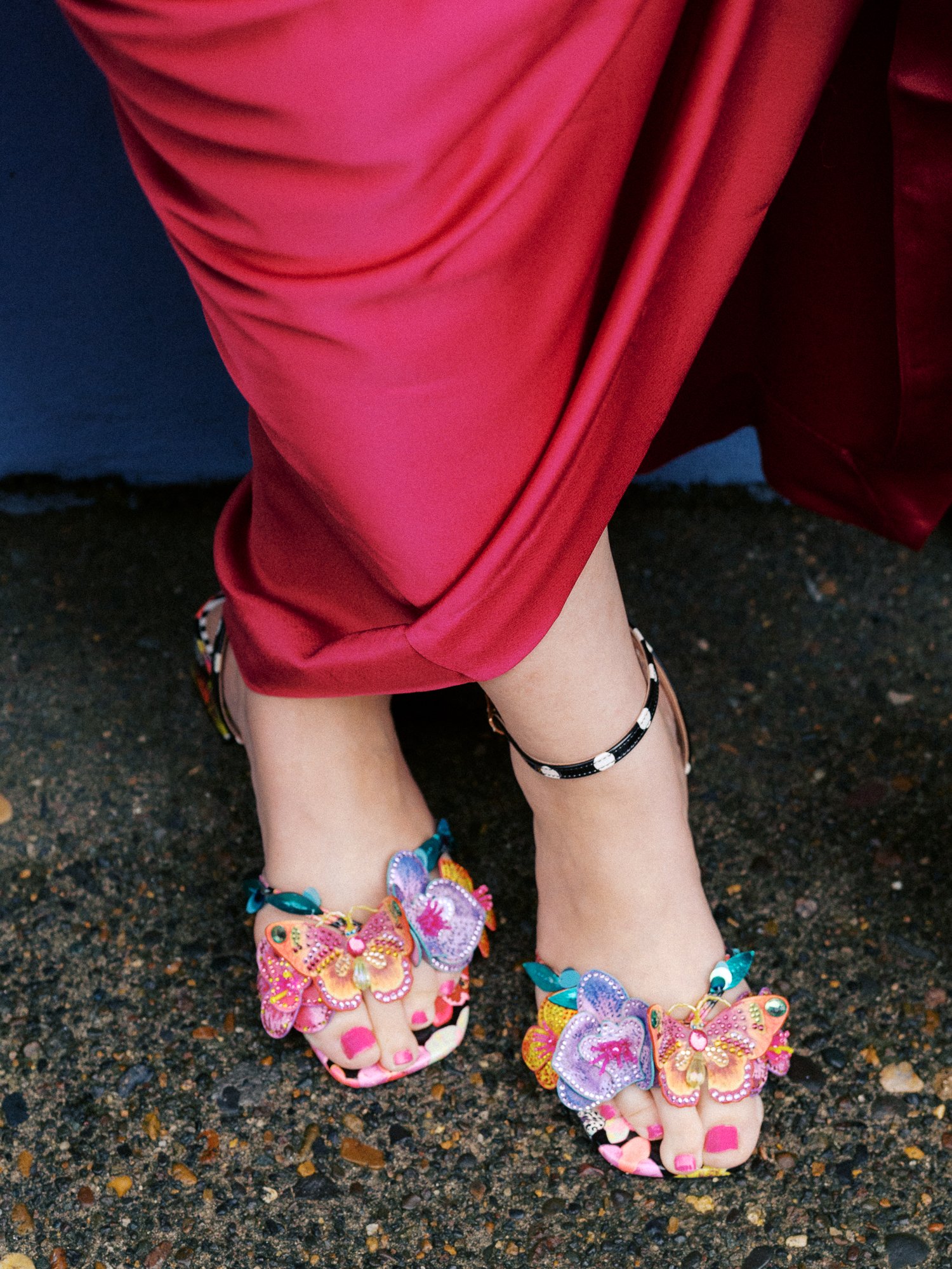 Colorful butterfly-embellished heels peek out beneath a bright pink wedding guest dress.