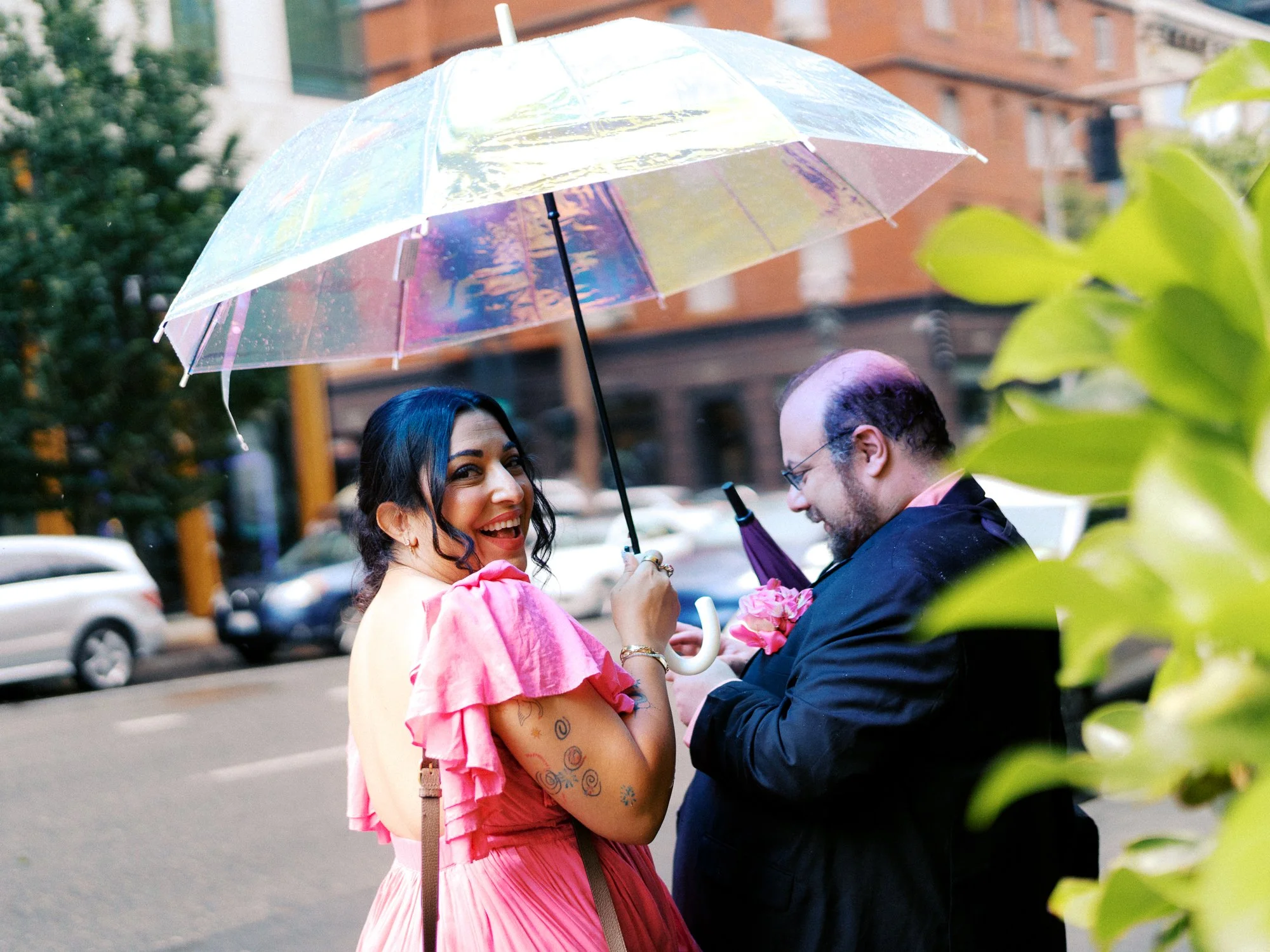 Wedding guest in a bright pink dress laughs beneath a clear umbrella on a rainy Portland street.