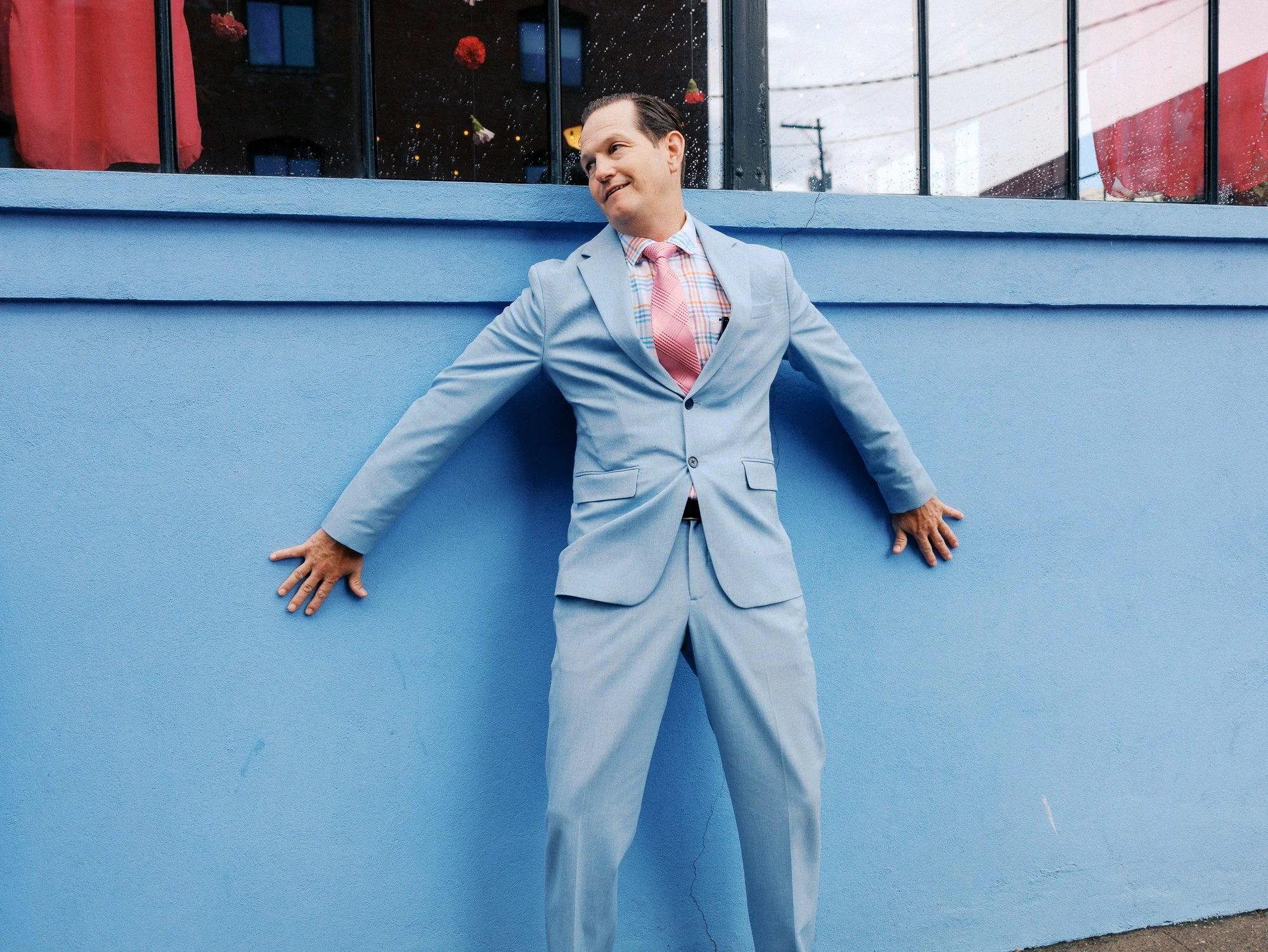 Wedding guest in a pale blue suit poses against the blue exterior wall at Castaway Portland.
