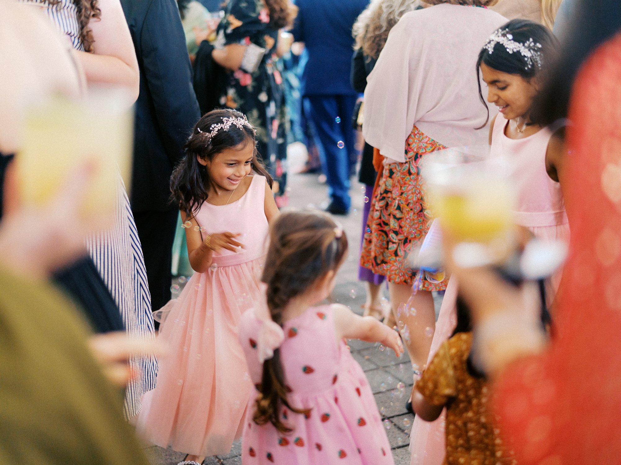 Children in pink dresses play with bubbles during the outdoor wedding celebration.
