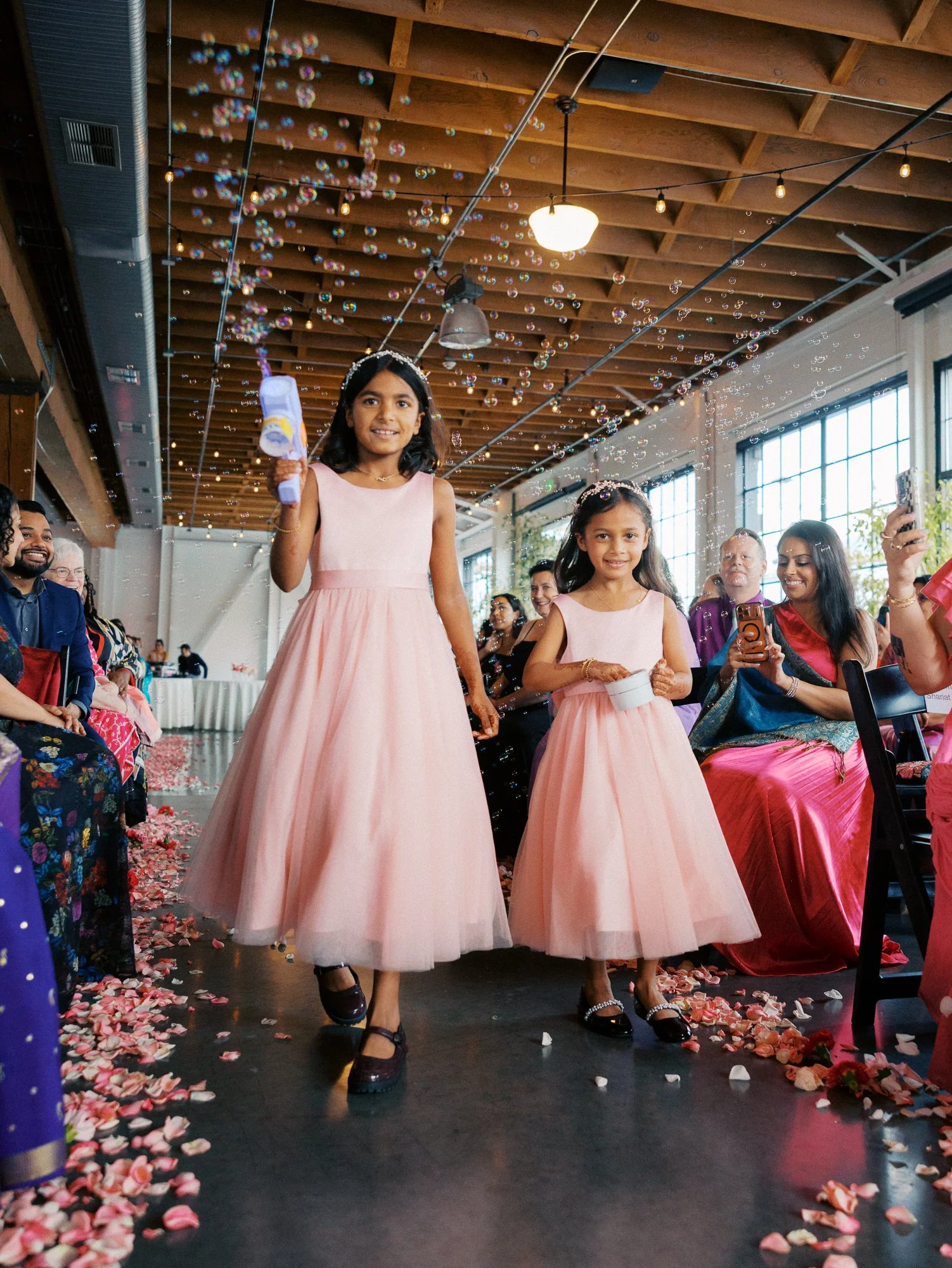 Two girls in matching pink dresses blow bubbles while walking down the ceremony aisle.