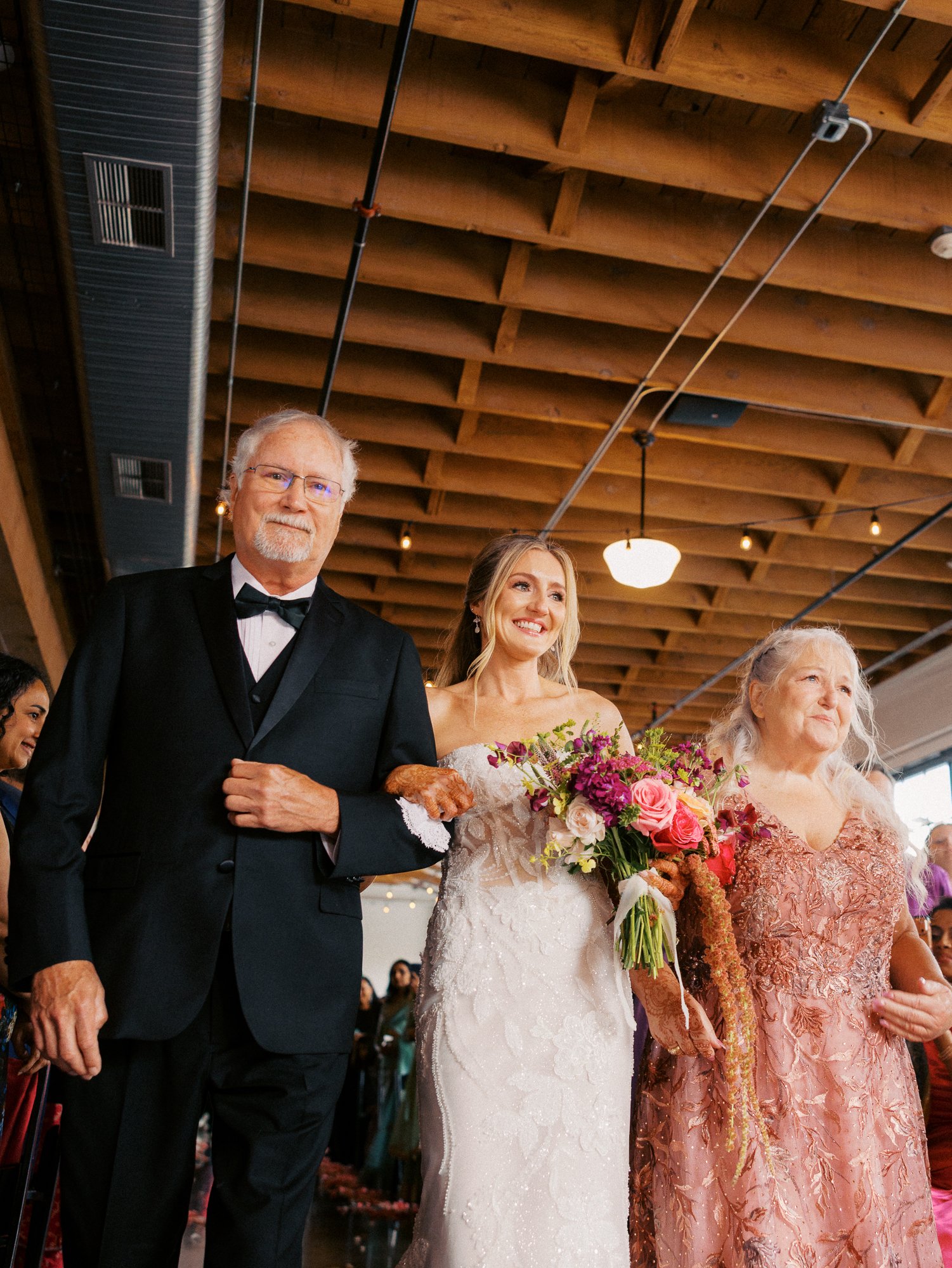 Bride walks down the aisle between two family members while holding a bright floral bouquet.