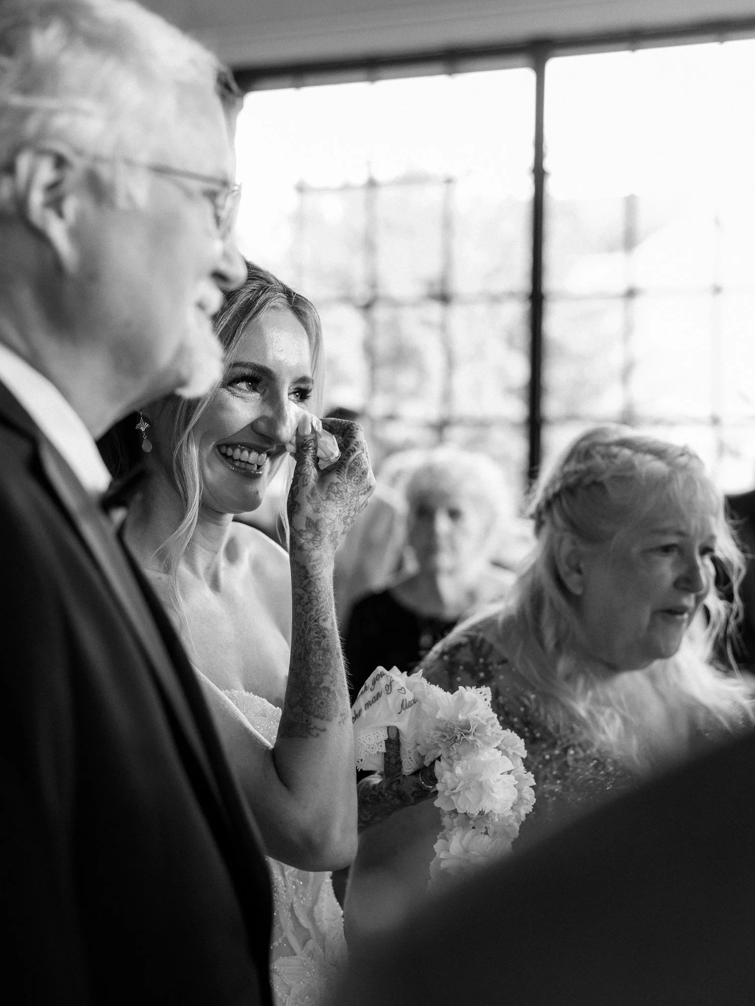 Black-and-white photo of the bride wiping tears while standing with family during the ceremony.