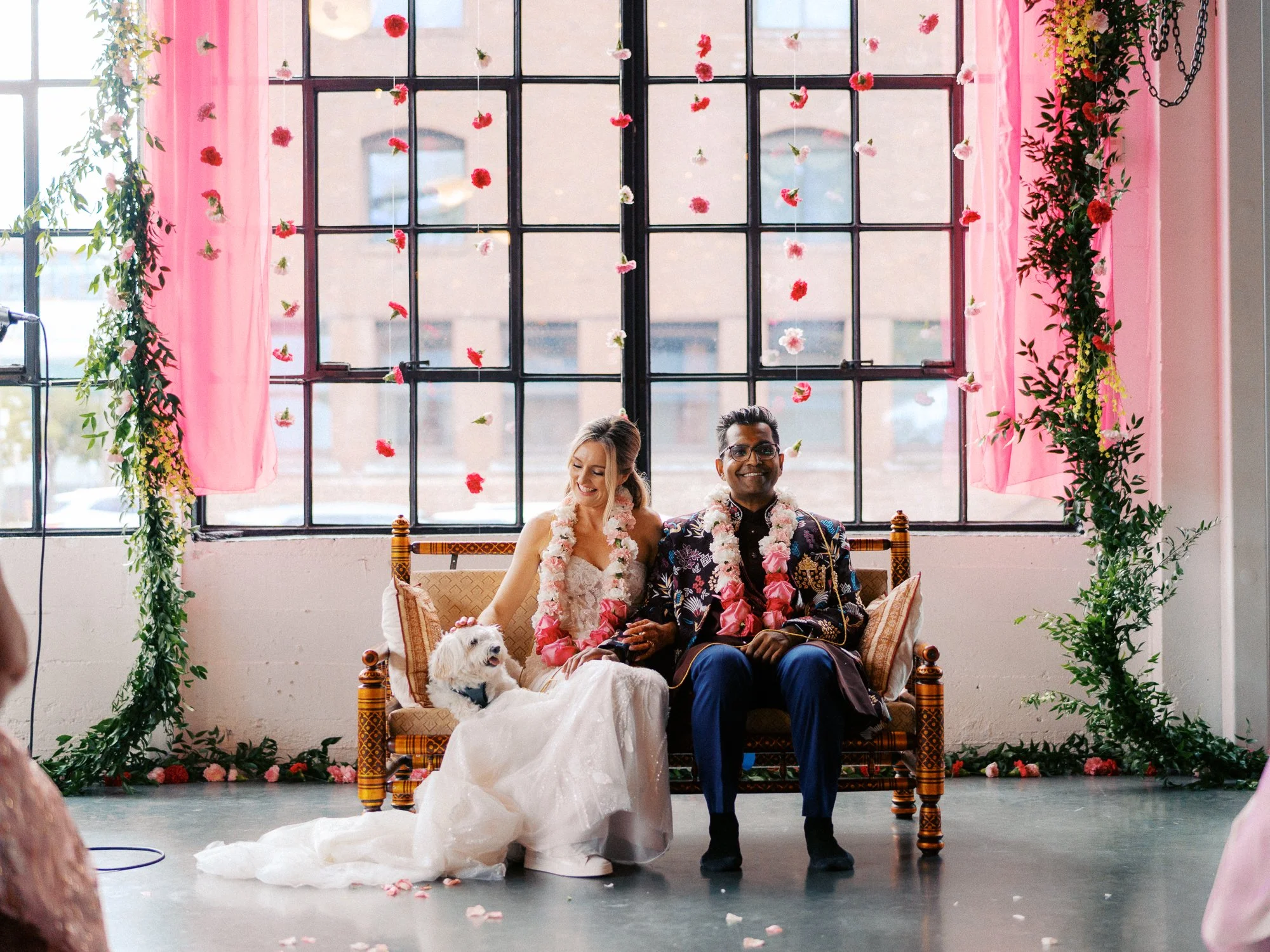 Bride and groom sit with their small white dog beneath suspended flowers and greenery.