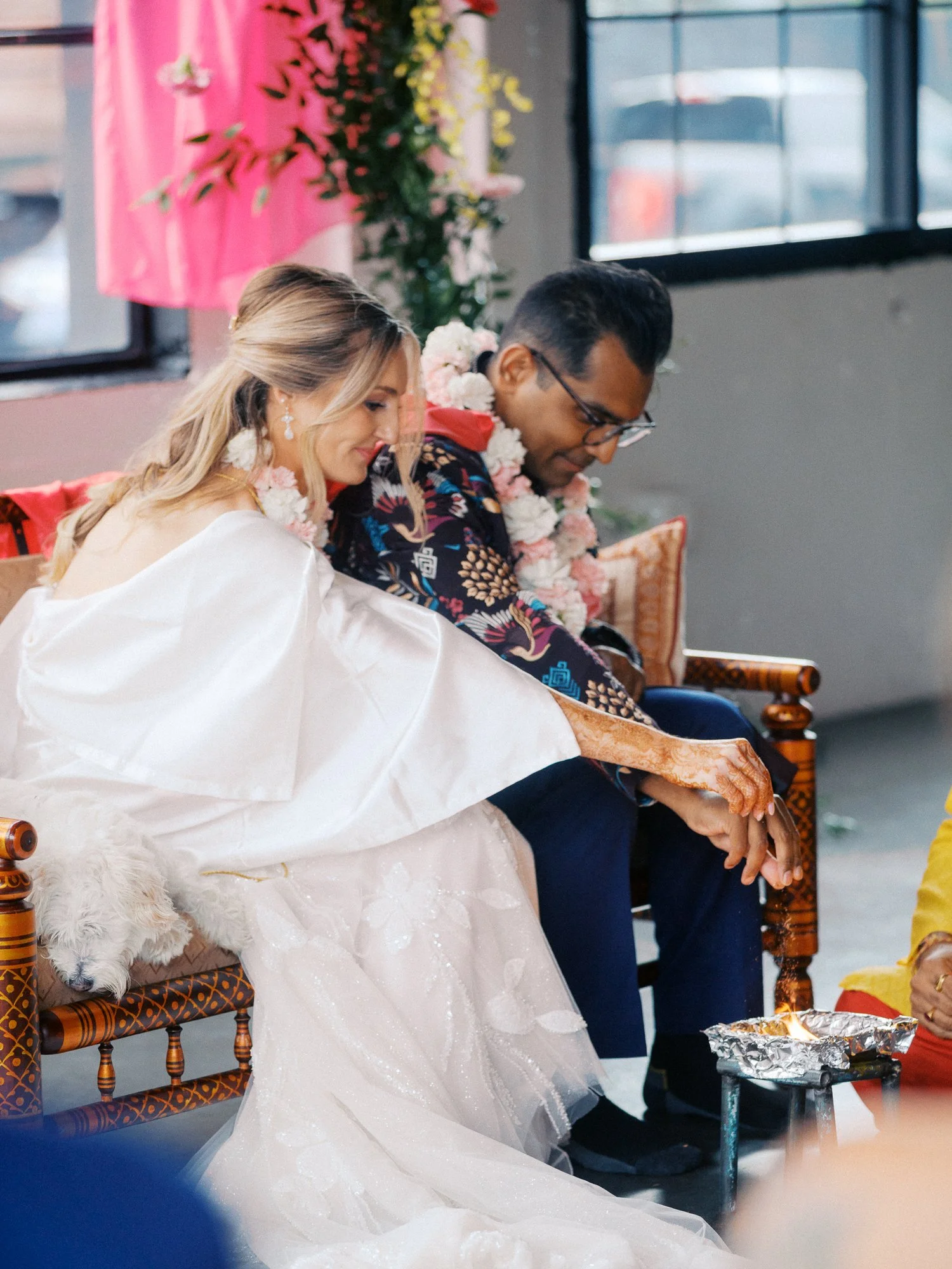Bride and groom hold hands during a seated wedding ritual, with their dog resting beside them.