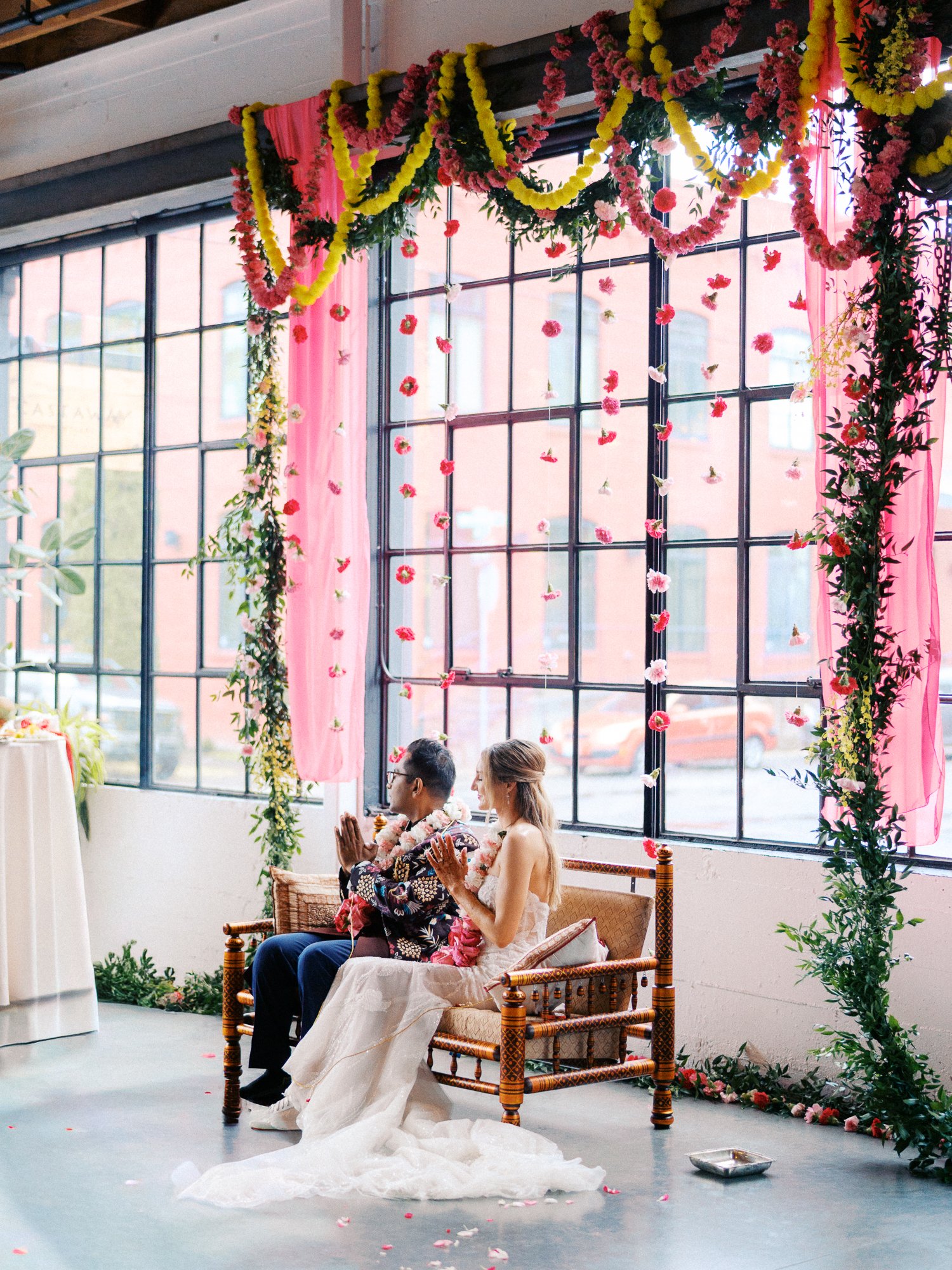 Bride and groom sit side by side with hands raised in prayer beneath the floral ceremony installation.