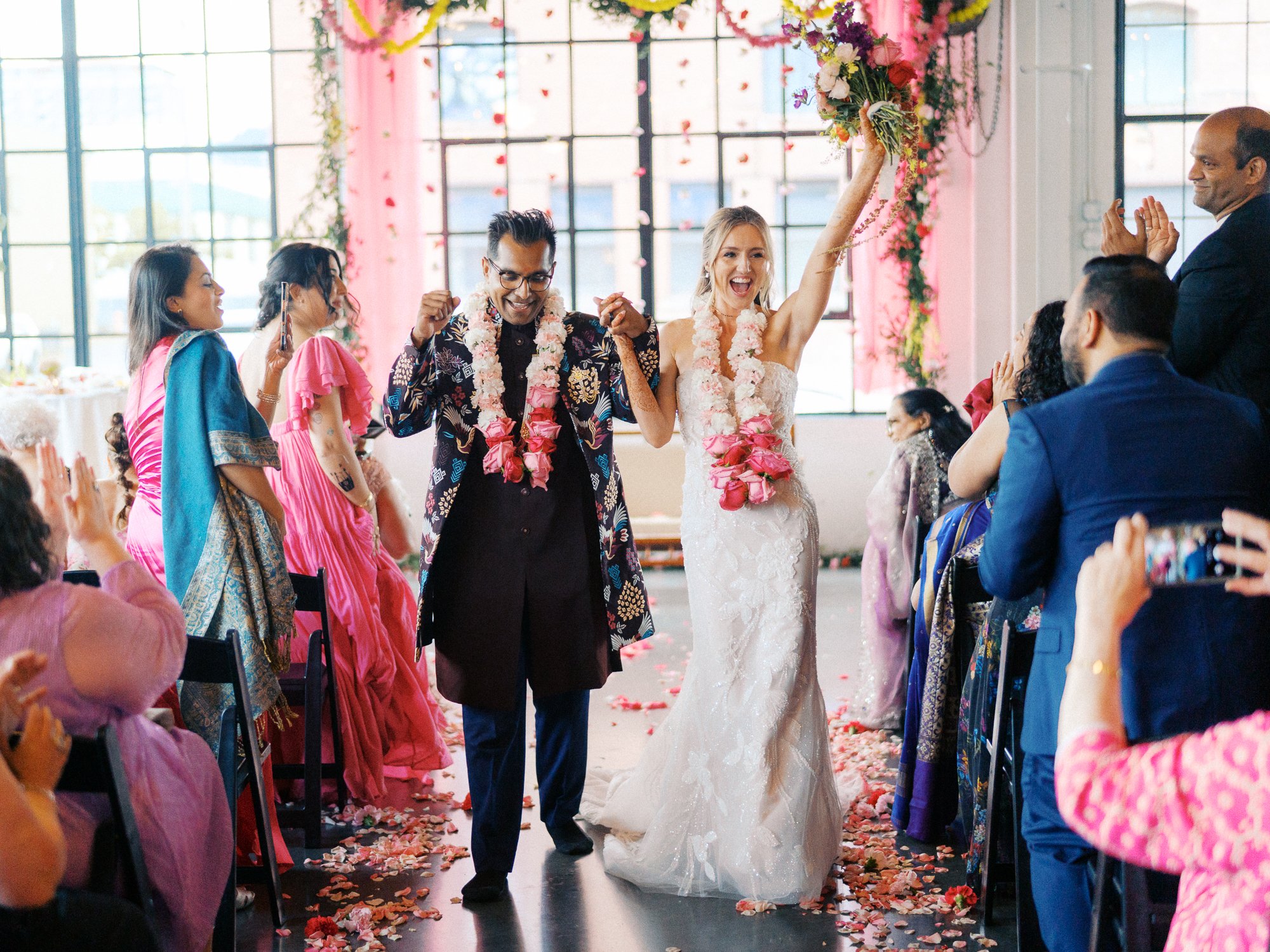 Bride raises her bouquet as she and the groom walk down the petal-covered aisle.