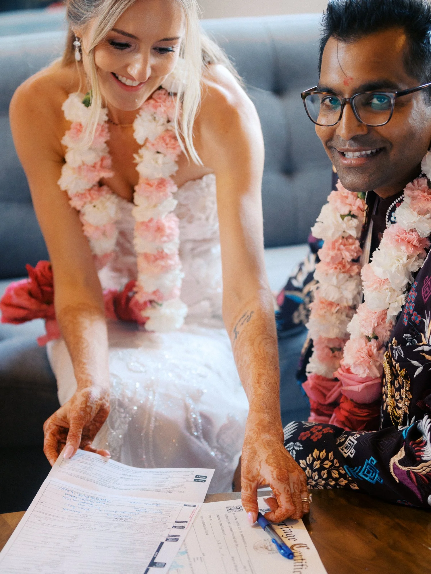 Bride and groom sign wedding documents while wearing pink and white floral garlands.