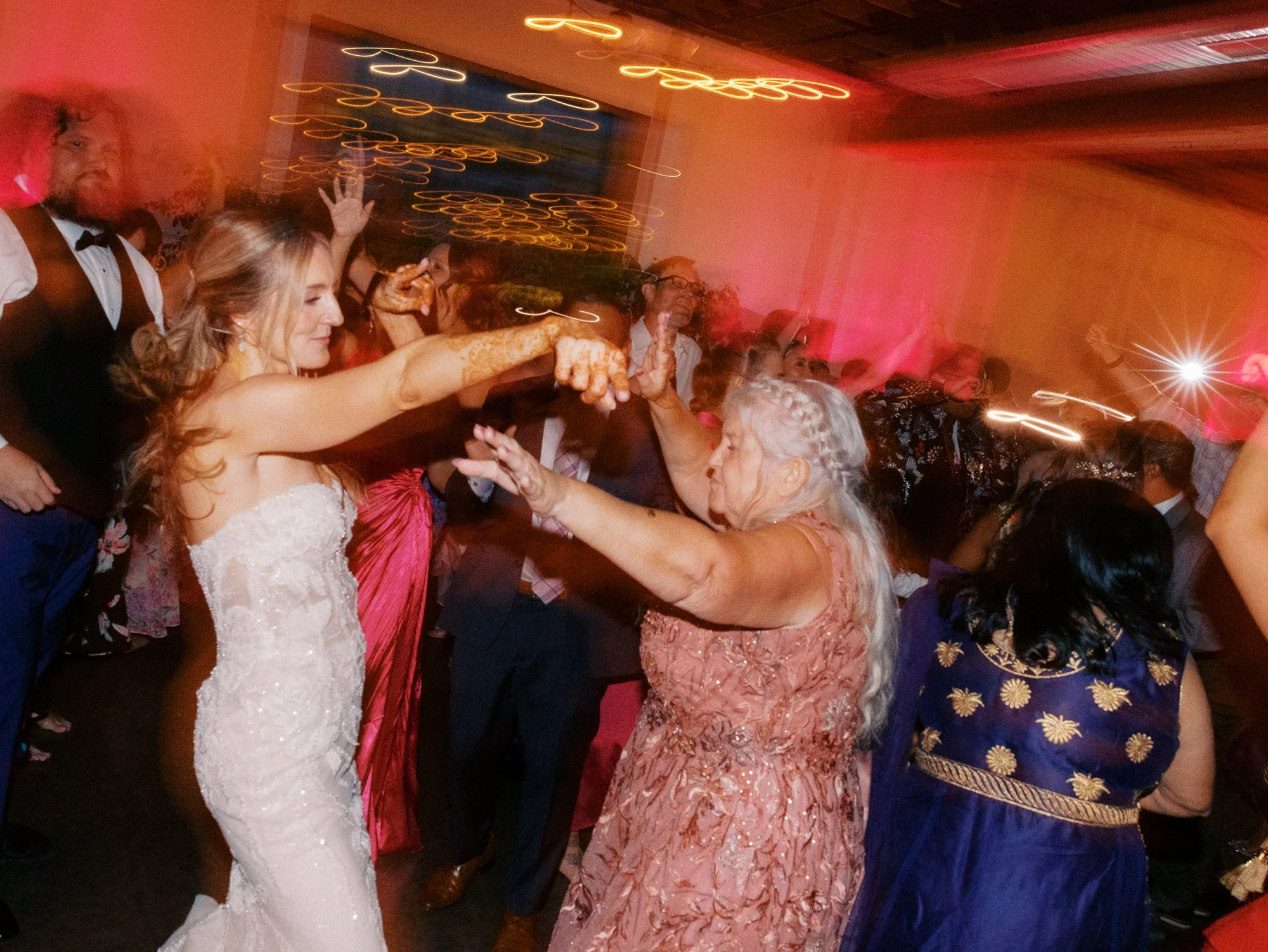 Bride dances with an older woman as guests fill the dance floor under warm reception lighting.