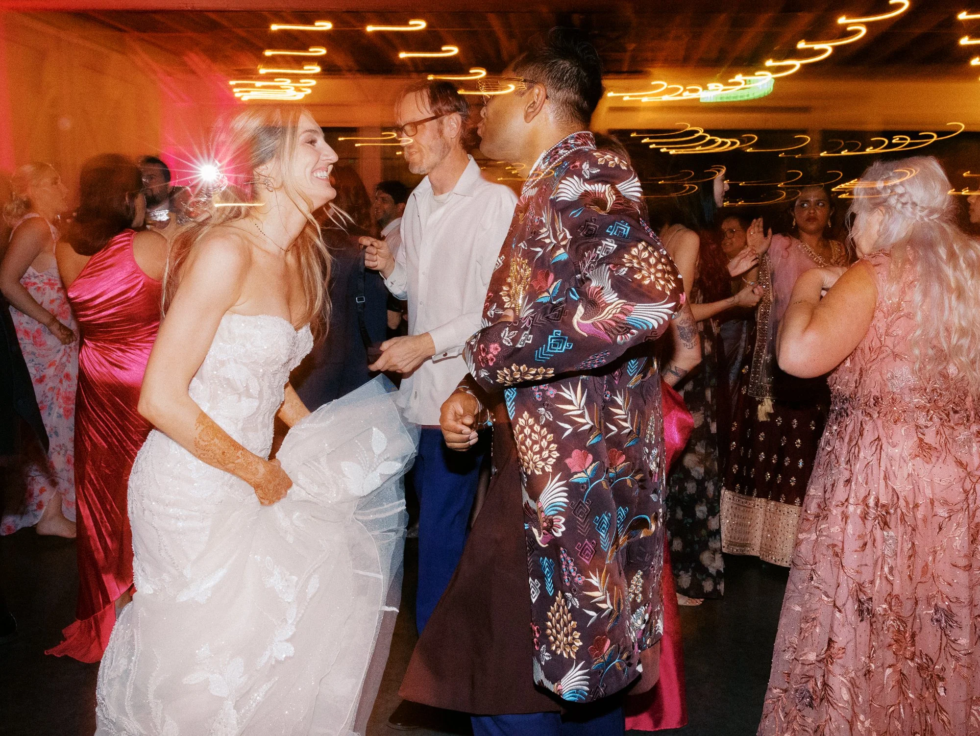 Bride and groom dance together surrounded by guests and glowing string lights.