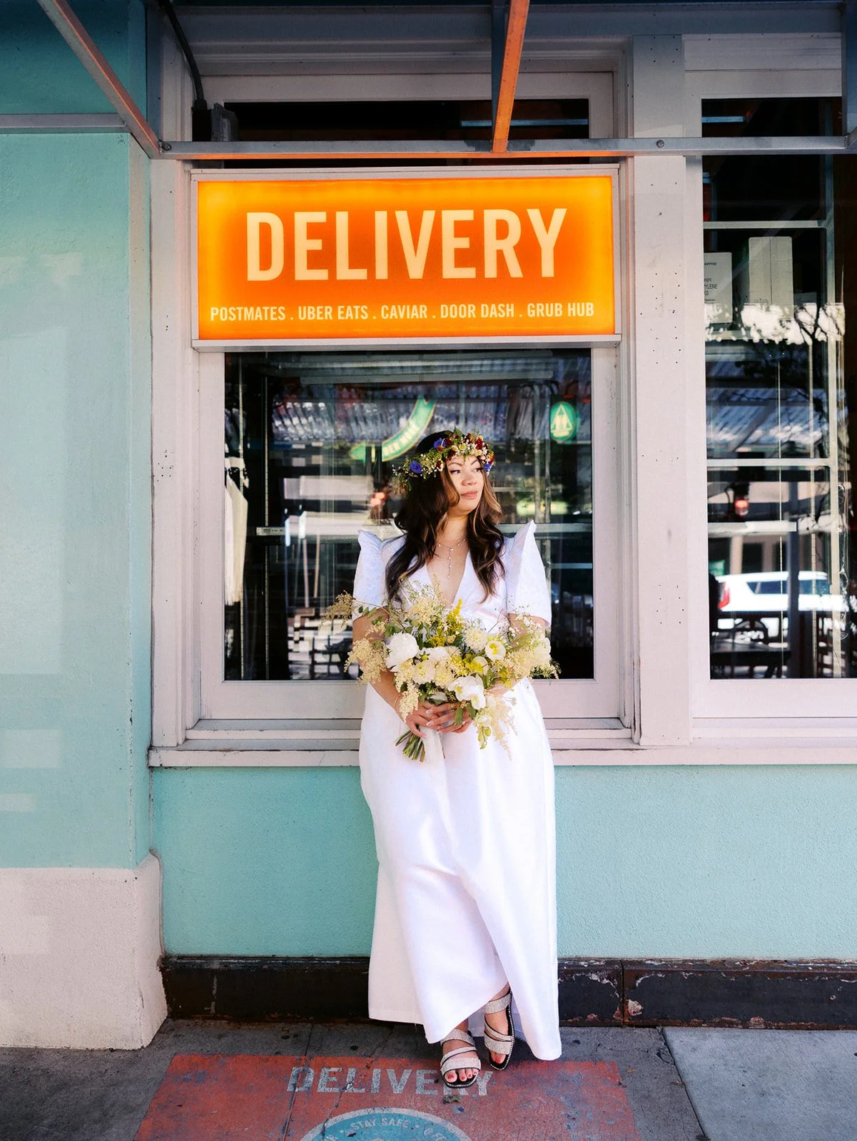Bride in a white puff-sleeve dress and colorful flower crown holds her bouquet outside a turquoise storefront in Portland.