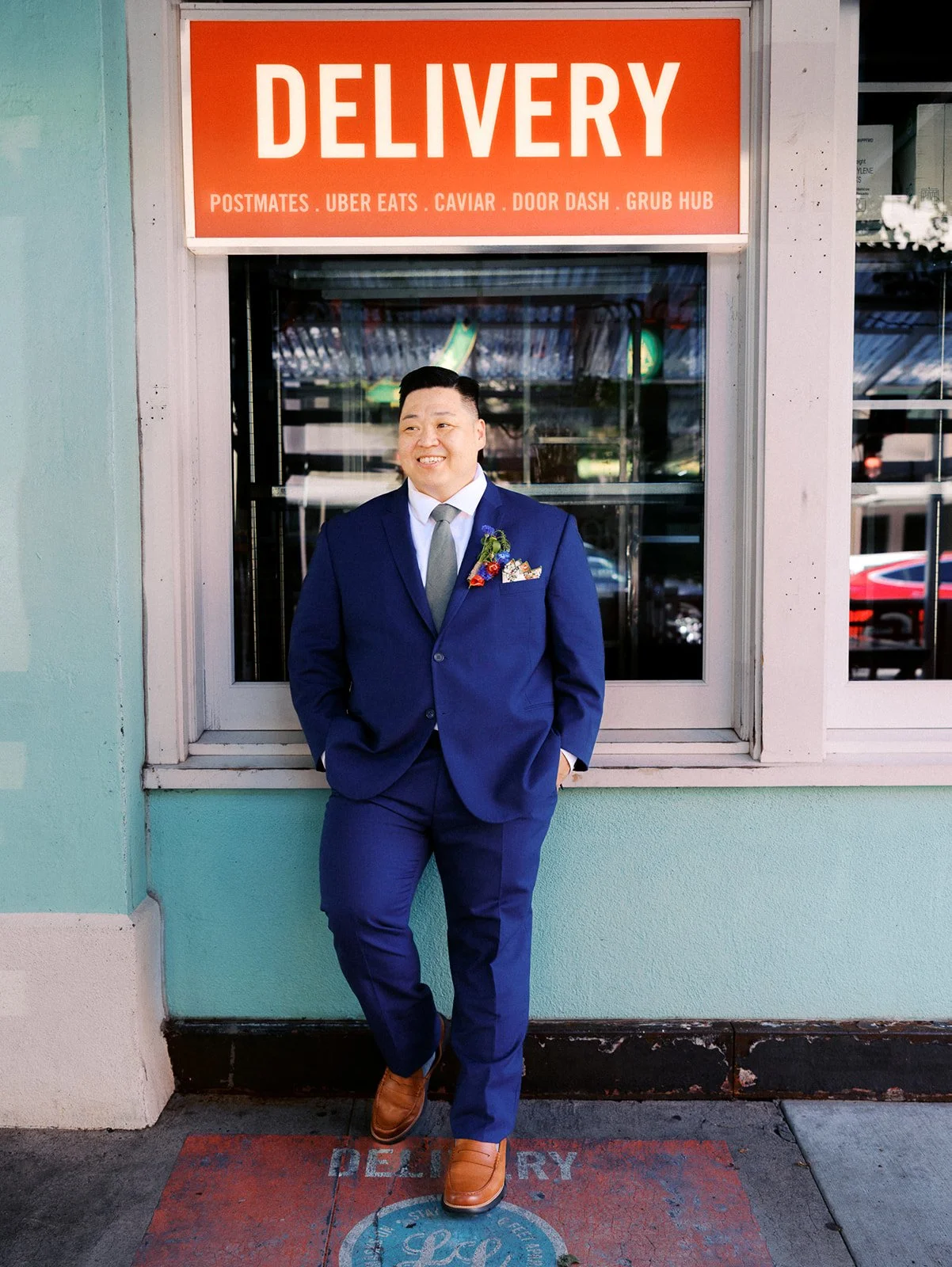 Groom in a navy blue suit and floral boutonniere smiles while leaning against a turquoise storefront beneath an orange sign.