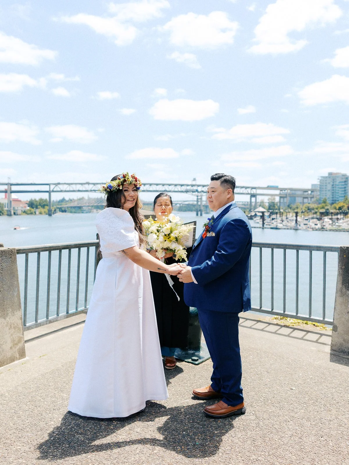 Couple holds hands during their waterfront elopement ceremony in Portland, with a bridge and river behind them.