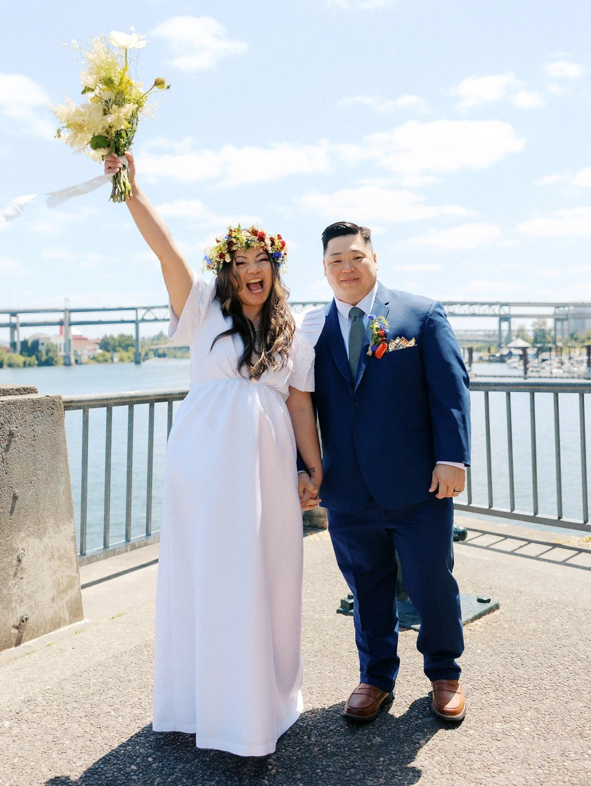 Bride raises her wedding bouquet overhead while laughing beside her partner in a navy suit after their ceremony.