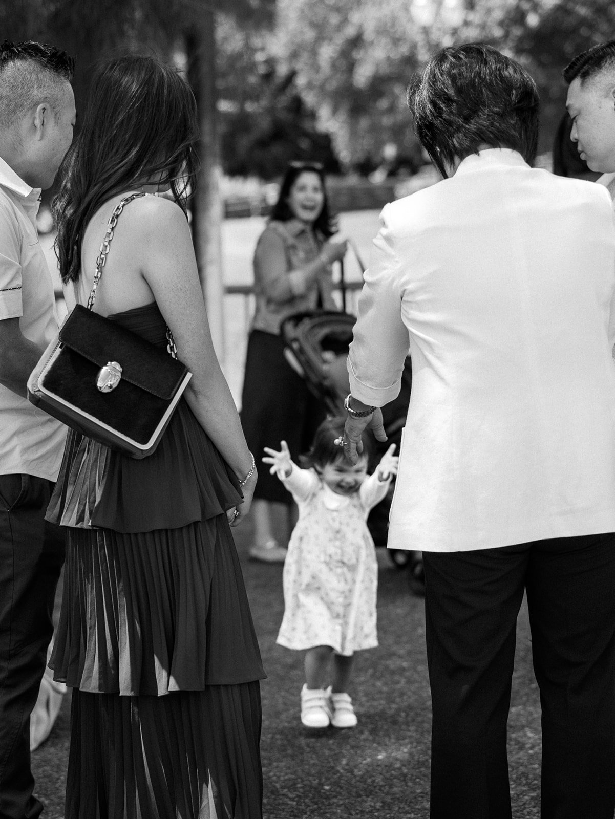 Black and white photograph of a toddler reaching toward wedding guests during an outdoor gathering.