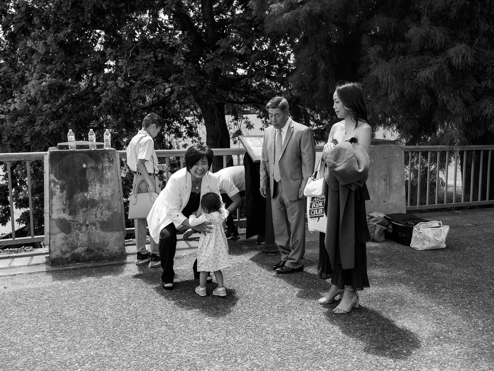 Black and white candid of wedding guests gathered around a small child near a tree-lined waterfront railing.