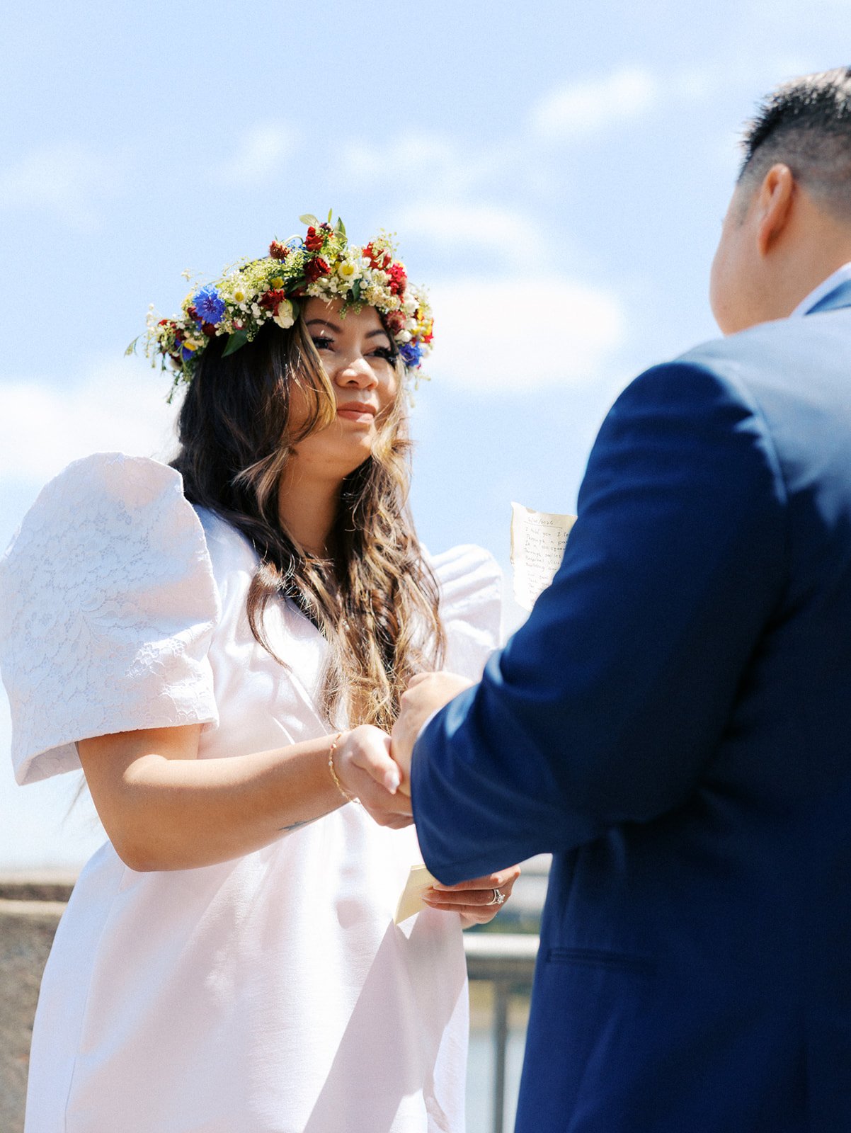 Bride wearing a colorful wildflower crown and puff-sleeve wedding dress holds her partner's hand during their vows.