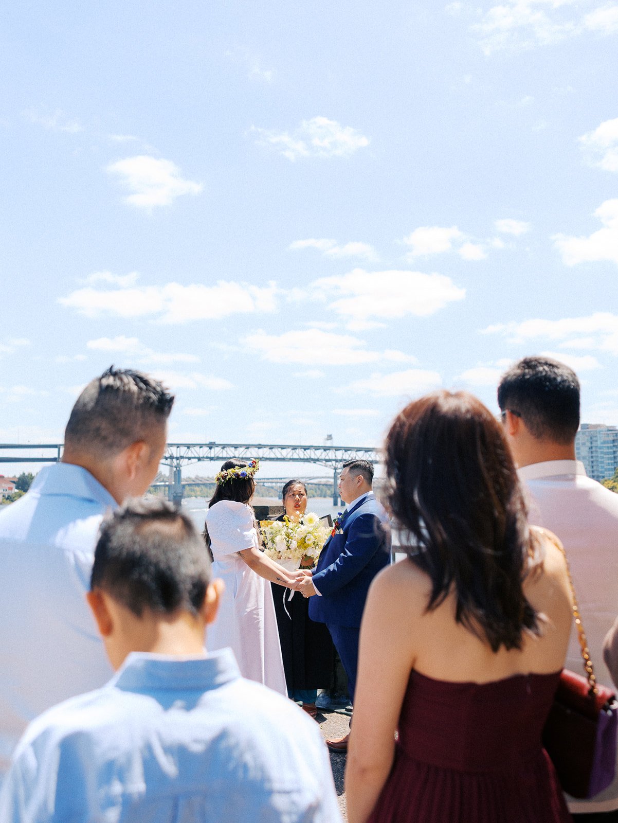 Guests watch from the foreground as a couple holds hands during their intimate riverside wedding ceremony.