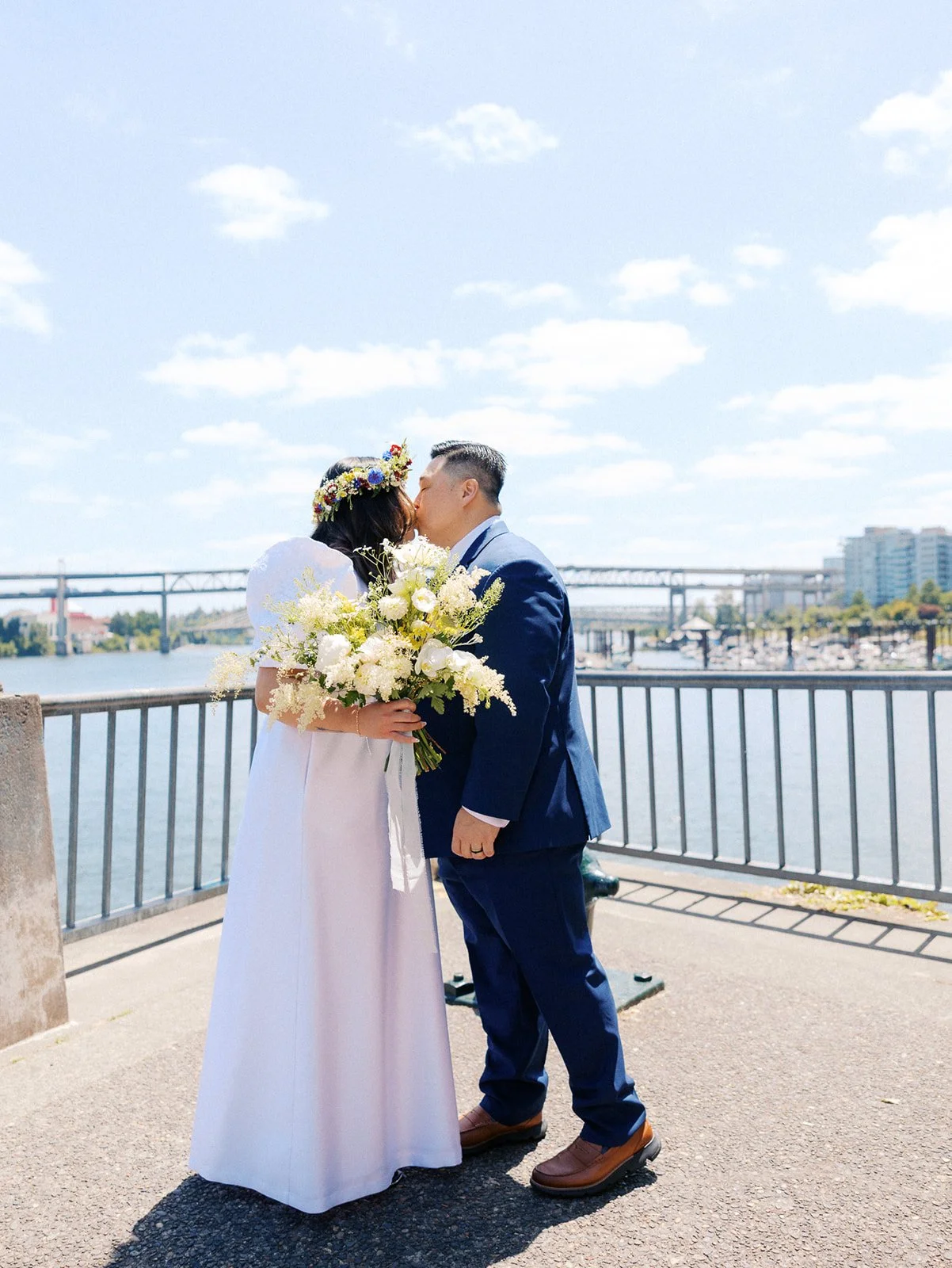 Newlyweds share a kiss beside the Portland waterfront, with the bride holding a bouquet of white flowers.
