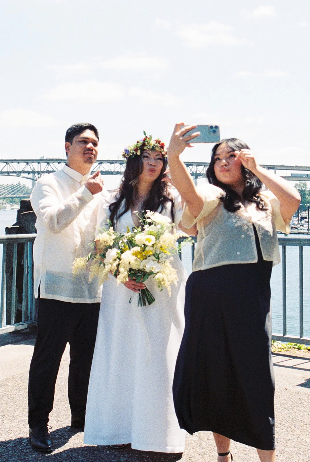 Bride wearing a flower crown poses for a waterfront selfie with two wedding guests beneath a sunny sky.