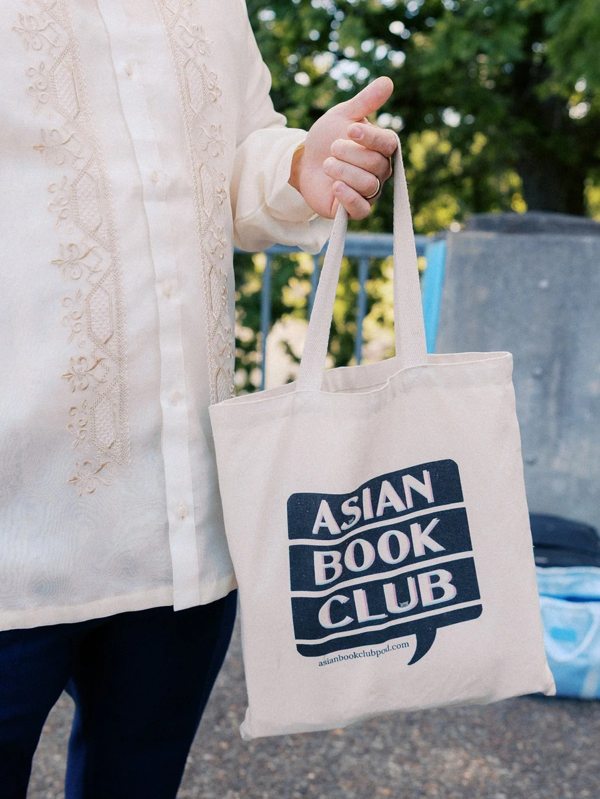 Close-up of a wedding guest in an embroidered cream shirt holding a canvas Asian Book Club tote bag.