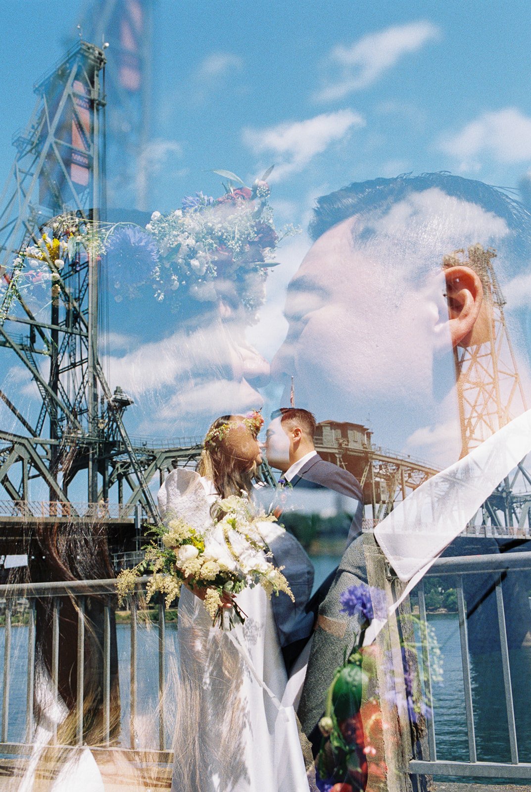 Artistic double exposure of newlyweds kissing, layered with a steel bridge, blue sky, and colorful wedding flowers.